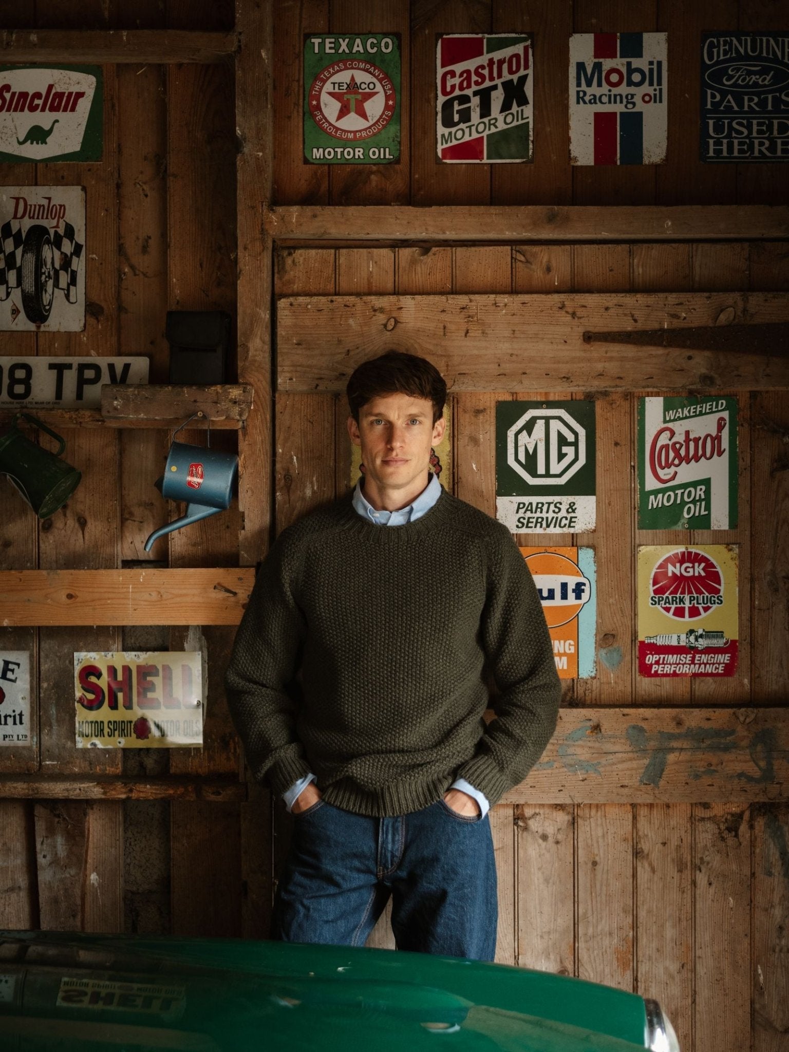 A man wearing a Campbell's of Beauly Moss Stitch Crew Neck Jumper and jeans stands in a wooden garage, hands in pockets, with vintage motor oil and car signs on the wall and part of a green car visible in the foreground.