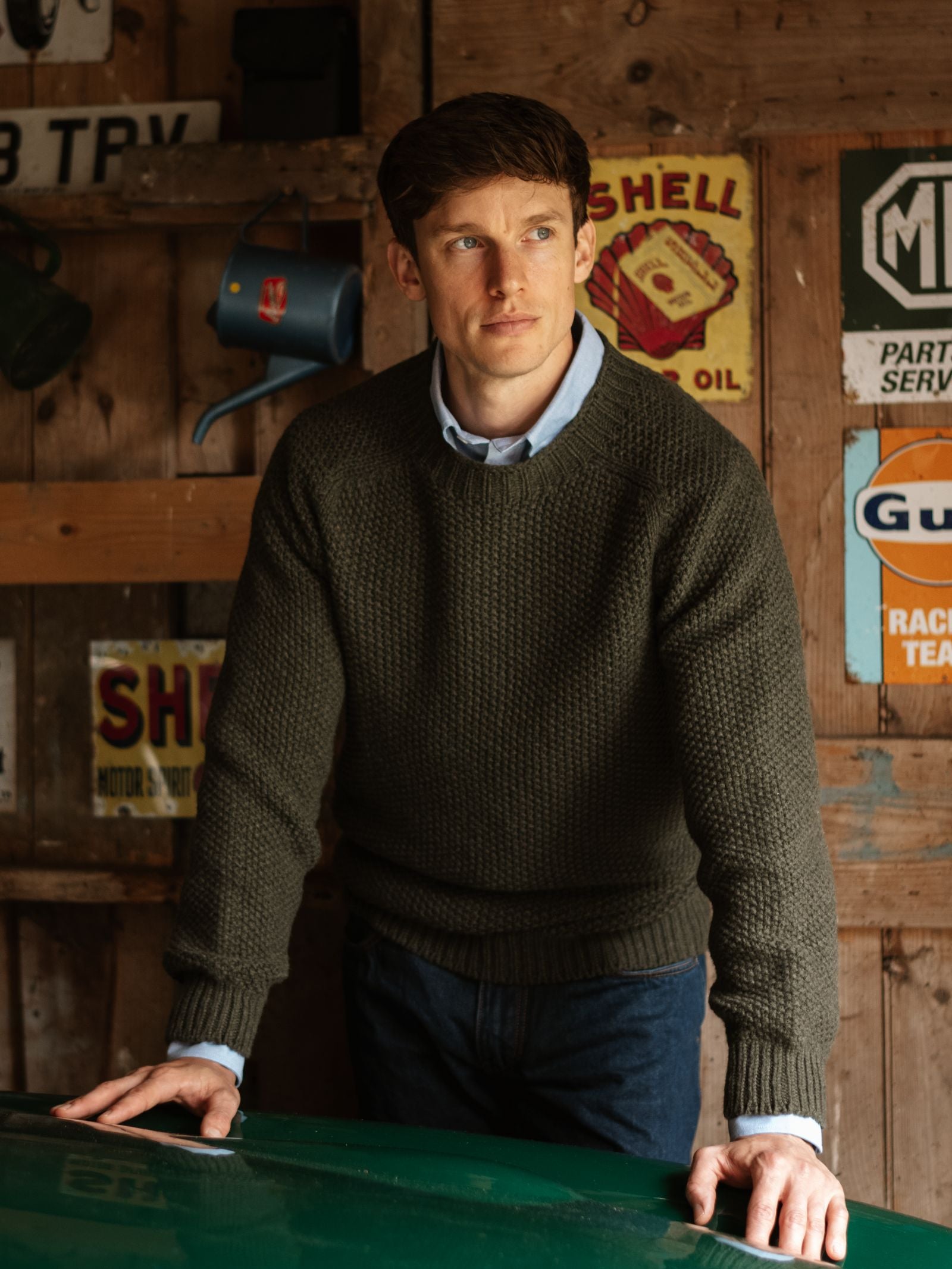 A man with short brown hair, wearing a loden Moss Stitch Crew Neck Jumper by Campbell's of Beauly over a blue collared shirt, stands leaning forward in a rustic wooden garage decorated with vintage signs and a watering can.