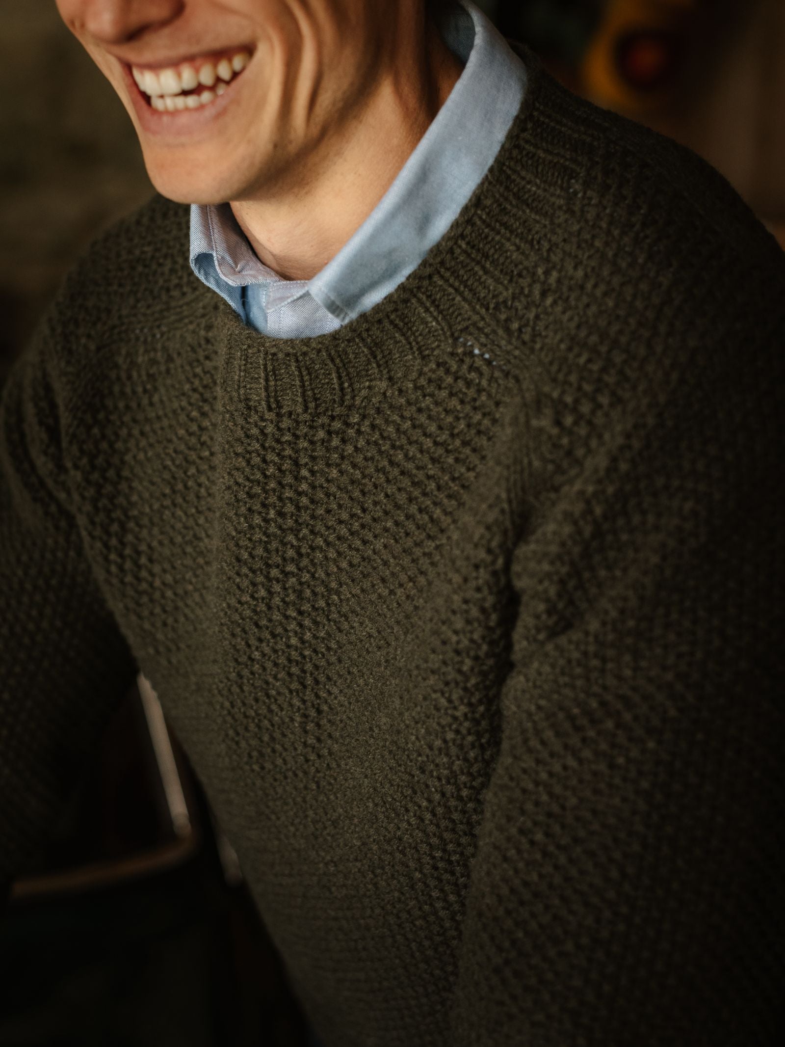 A person smiles, wearing a textured dark green Campbell's of Beauly Moss Stitch Crew Neck Jumper over a light blue collared shirt. The image is cropped to show just the lower face and upper torso.