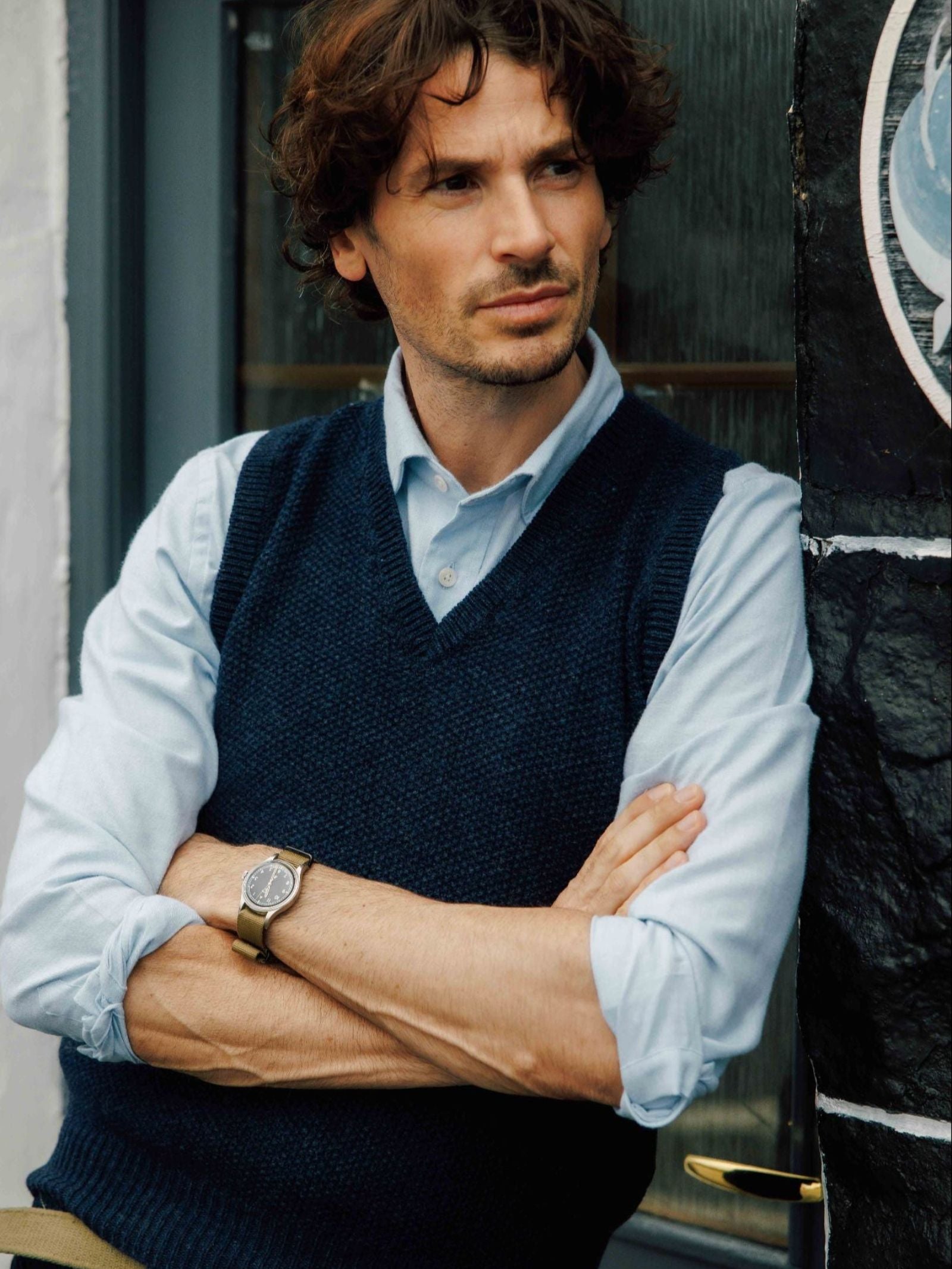 A man with wavy brown hair and light stubble stands with arms crossed, wearing a light blue shirt and the Campbell’s of Beauly Moss Stitch Knitted Slipover, made in the UK, paired with a wristwatch, looking thoughtfully to the side.