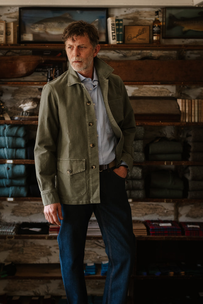 A man wearing a khaki green cotton moleskin Nevis Jacket by Campbell’s of Beauly with a button front and large patch pockets over a light blue shirt and dark jeans.