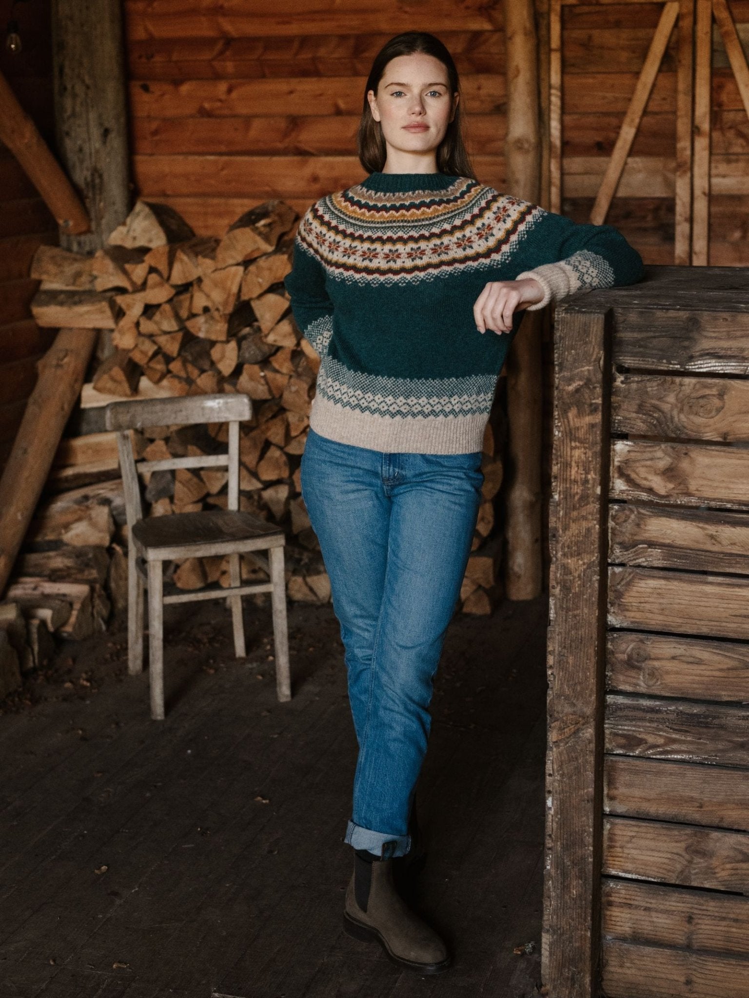 A woman in the Campbell's of Beauly Nordic Fairisle Crew Jumper, jeans, and boots leans against a wooden wall inside a rustic cabin, with stacked firewood and a wooden chair in the background.