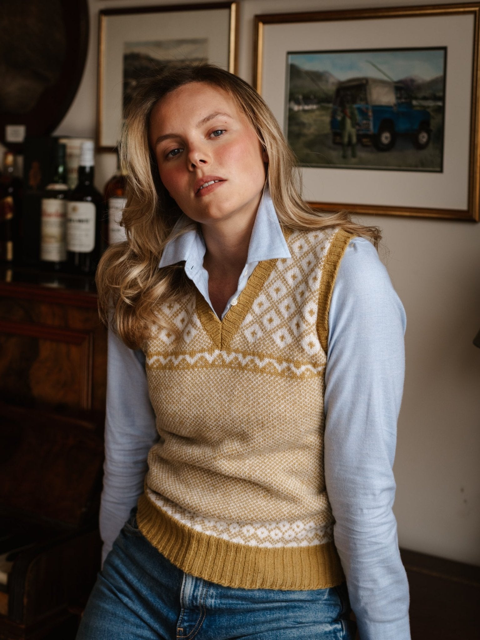 A woman with blonde hair wears the Campbell's of Beauly Auld Stock Nordic V Tank, a yellow and white Fair Isle sweater vest, over a light blue collared shirt. She poses indoors in front of framed paintings and bottles on a wooden shelf.