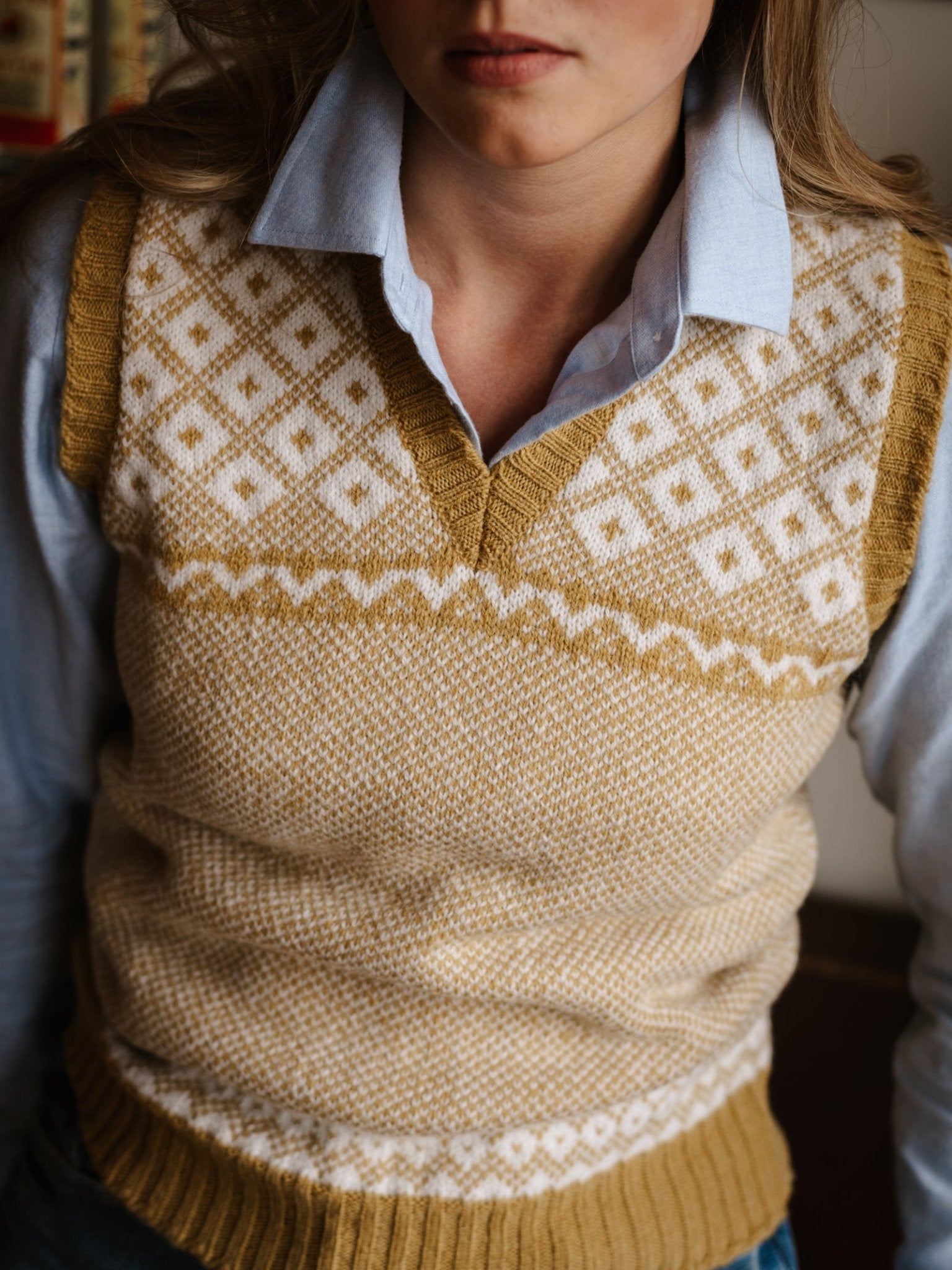 A person wears the Campbell's of Beauly Auld Stock Nordic V Tank, a mustard yellow and white Fair Isle sweater vest, over a light blue collared shirt. Their face is partially visible, with long light brown hair shown shoulders to waist.