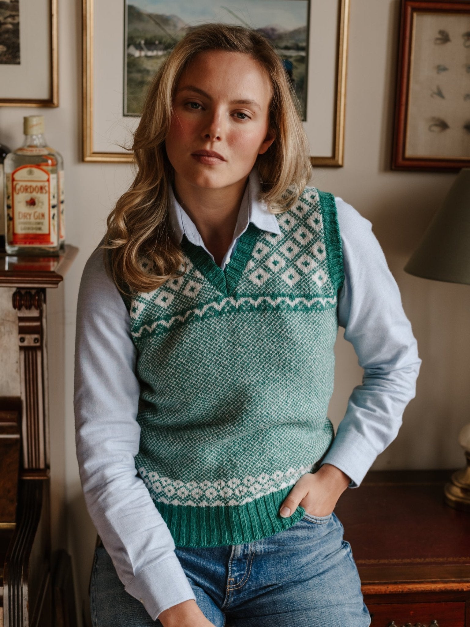 A person with long, wavy blond hair wears the Campbell's of Beauly Auld Stock Nordic V Tank, a green and white Fair Isle sweater vest, over a light blue shirt and jeans, posing indoors next to a cabinet with gin and framed artwork.