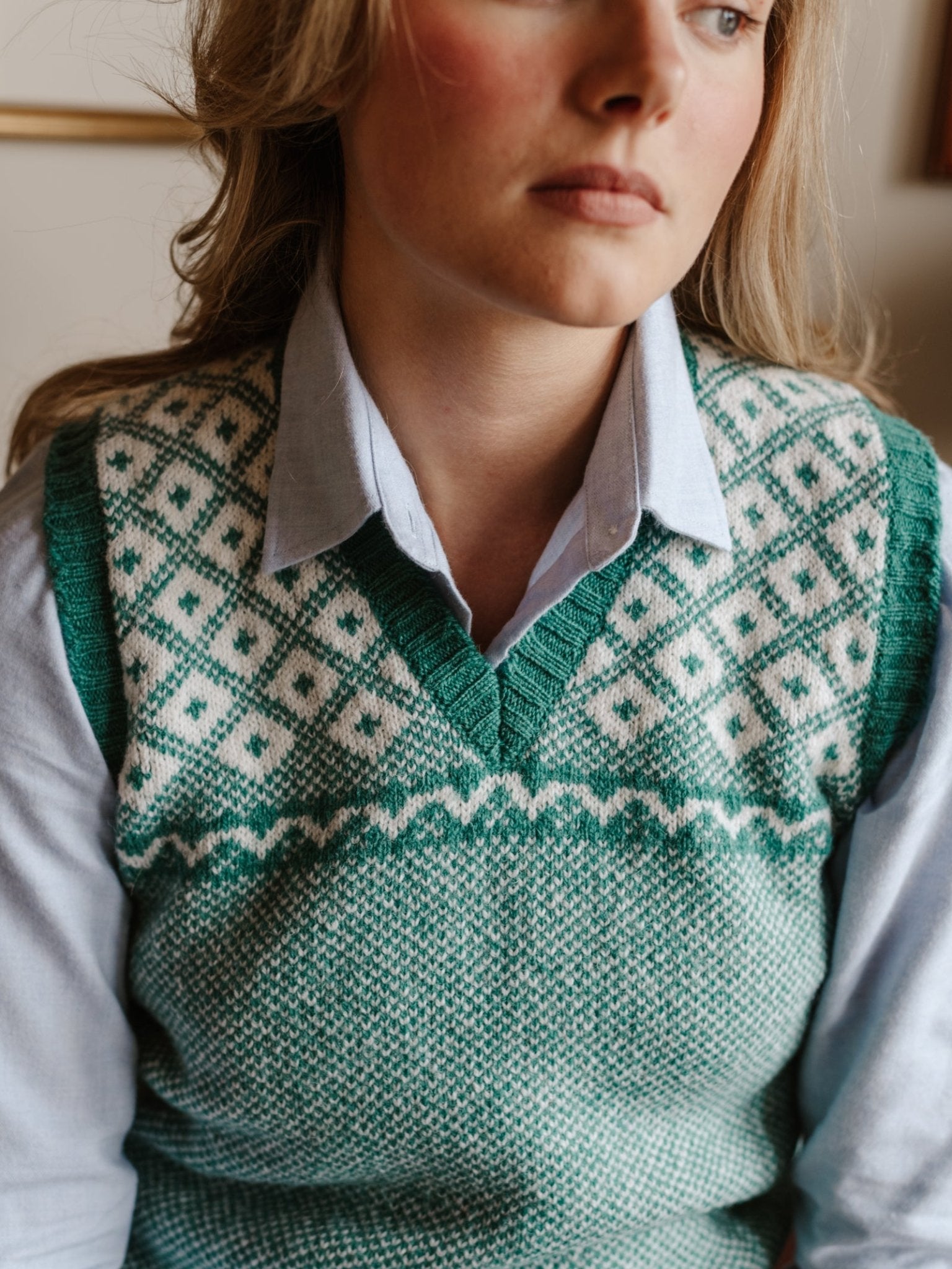 A woman in a light blue collared shirt and the Campbell's of Beauly Auld Stock Nordic V Tank—a green and white Fair Isle slipover with diamond patterns—looks slightly to the side with a neutral expression.