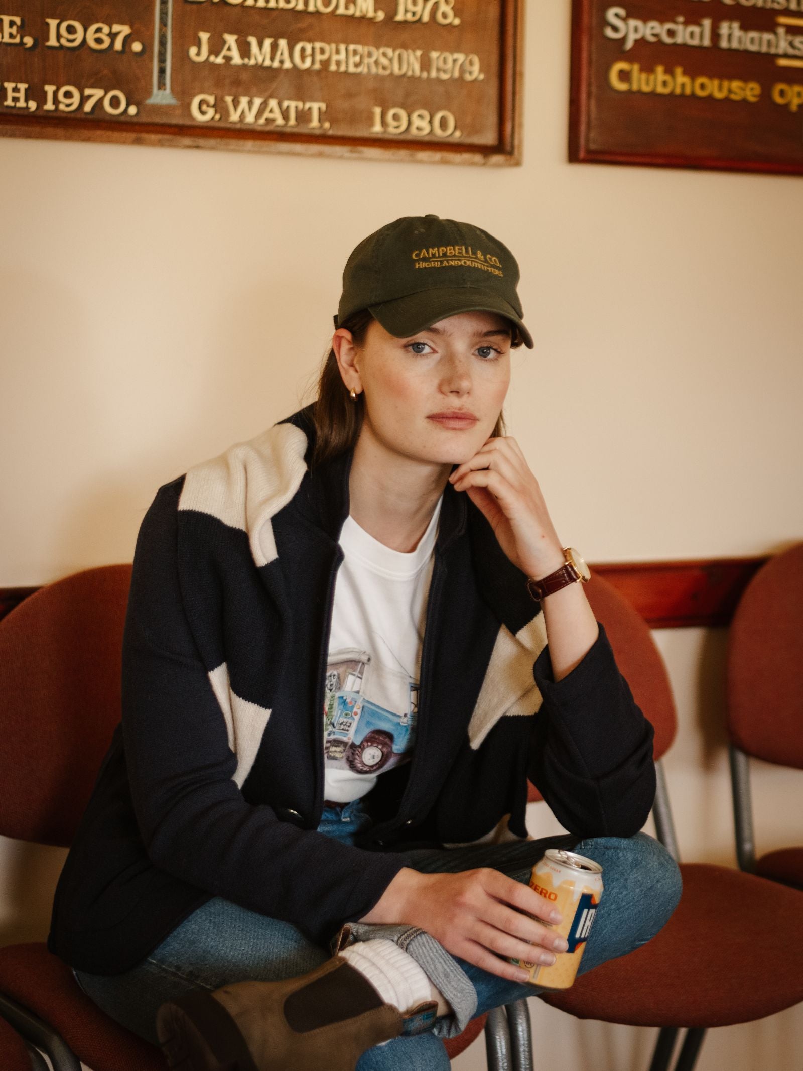 A woman in classic everyday wear—a navy jacket, white shirt, jeans, and the Unisex Campbell's Baseball Cap by Campbell's of Beauly—sits on a red chair holding a can, with plaques and brown chairs in the background.