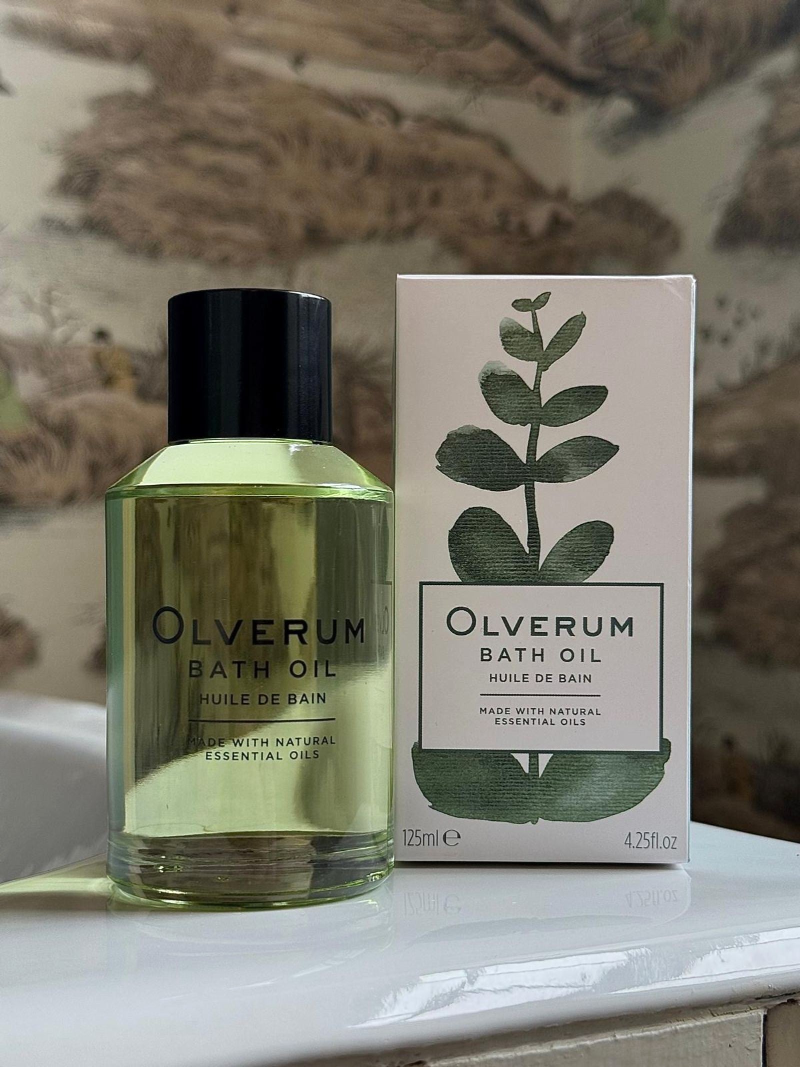 A bottle of OLVERUM Bath Oil stands beside its matching box with a green plant design. Both sit on a white surface, set against nature-inspired wallpaper—ideal for a soothing bath with luxurious essential oils.
