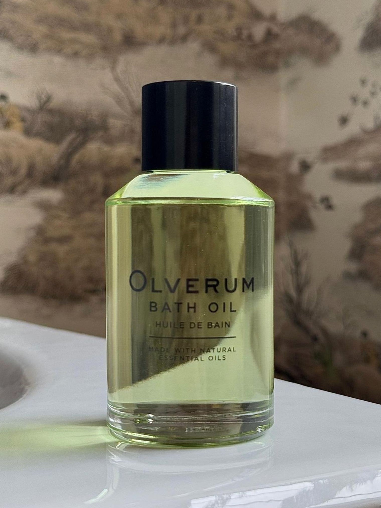 A clear OLVERUM Bath Oil bottle with a black cap sits on a white surface against a blurred, nature-themed background. Its label showcases natural essential oils, ideal for relaxing bath experiences.