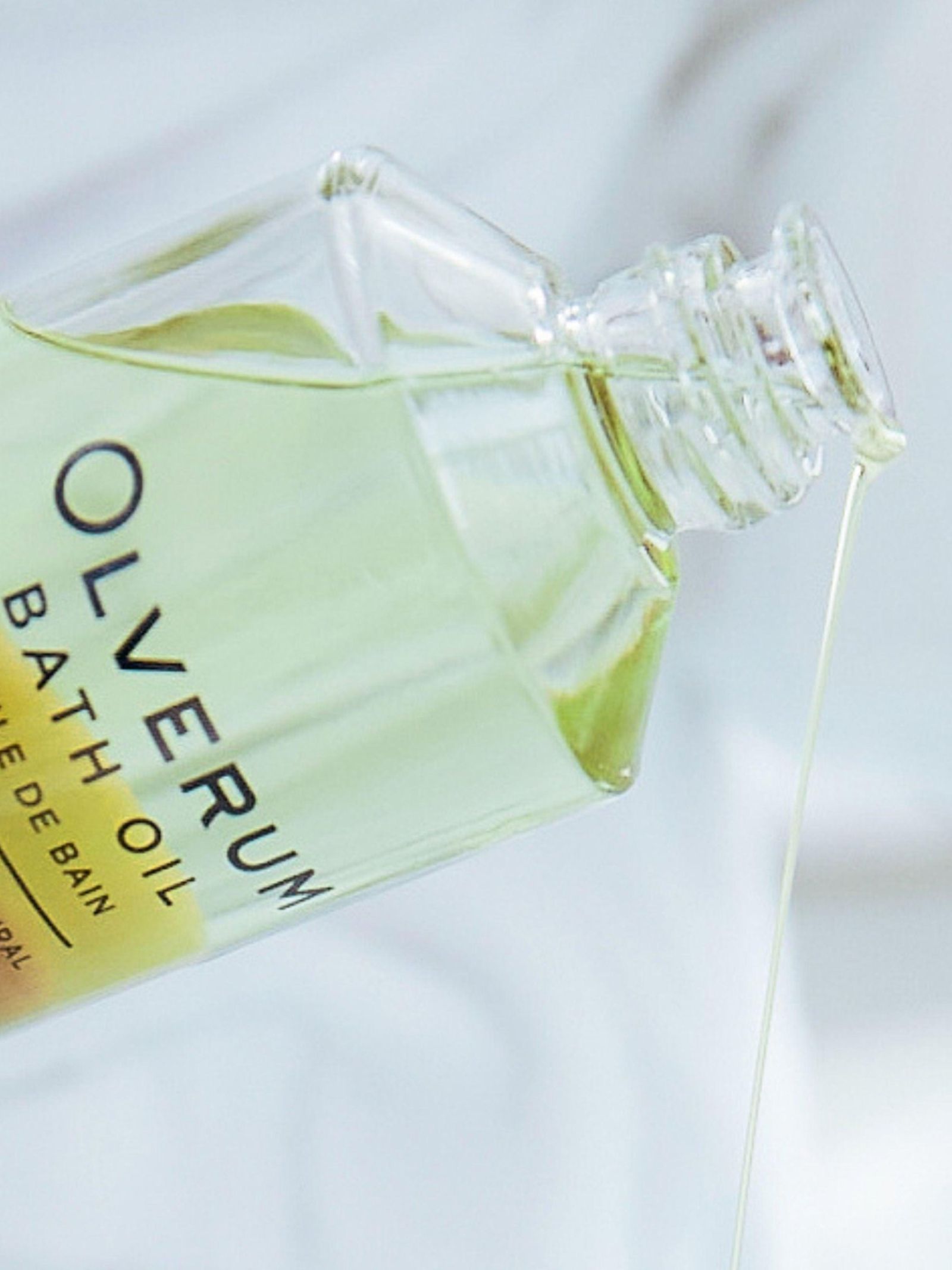 A close-up of a tilted OLVERUM Bath Oil bottle pours pale yellow, essential oil-infused bath oil against a soft, blurred background.