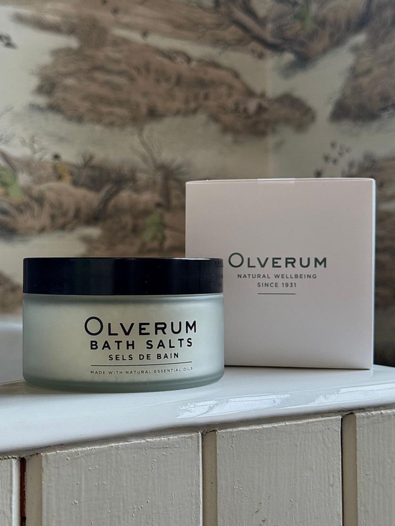 An OLVERUM Bath Salts jar with a black lid sits next to its matching box on a white surface, set against nature-inspired wallpaper that reflects the calming blend of essential oils and Dead Sea salt.