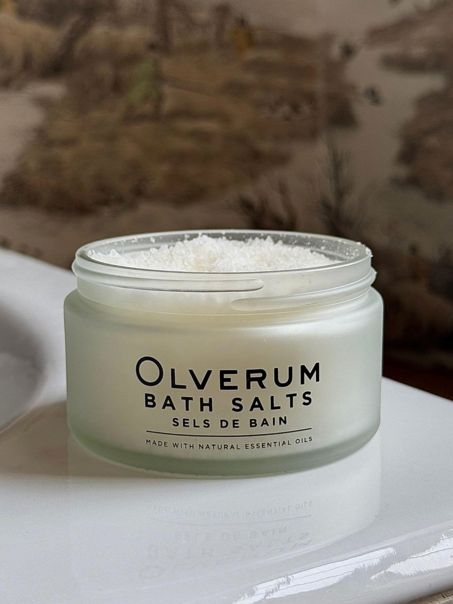 A frosted glass jar of OLVERUM Bath Salts, filled with coarse Dead Sea salt and natural essential oils, sits on a white surface. The label reads "OLVERUM Bath Salts Sels de Bain" with a blurred background in view.