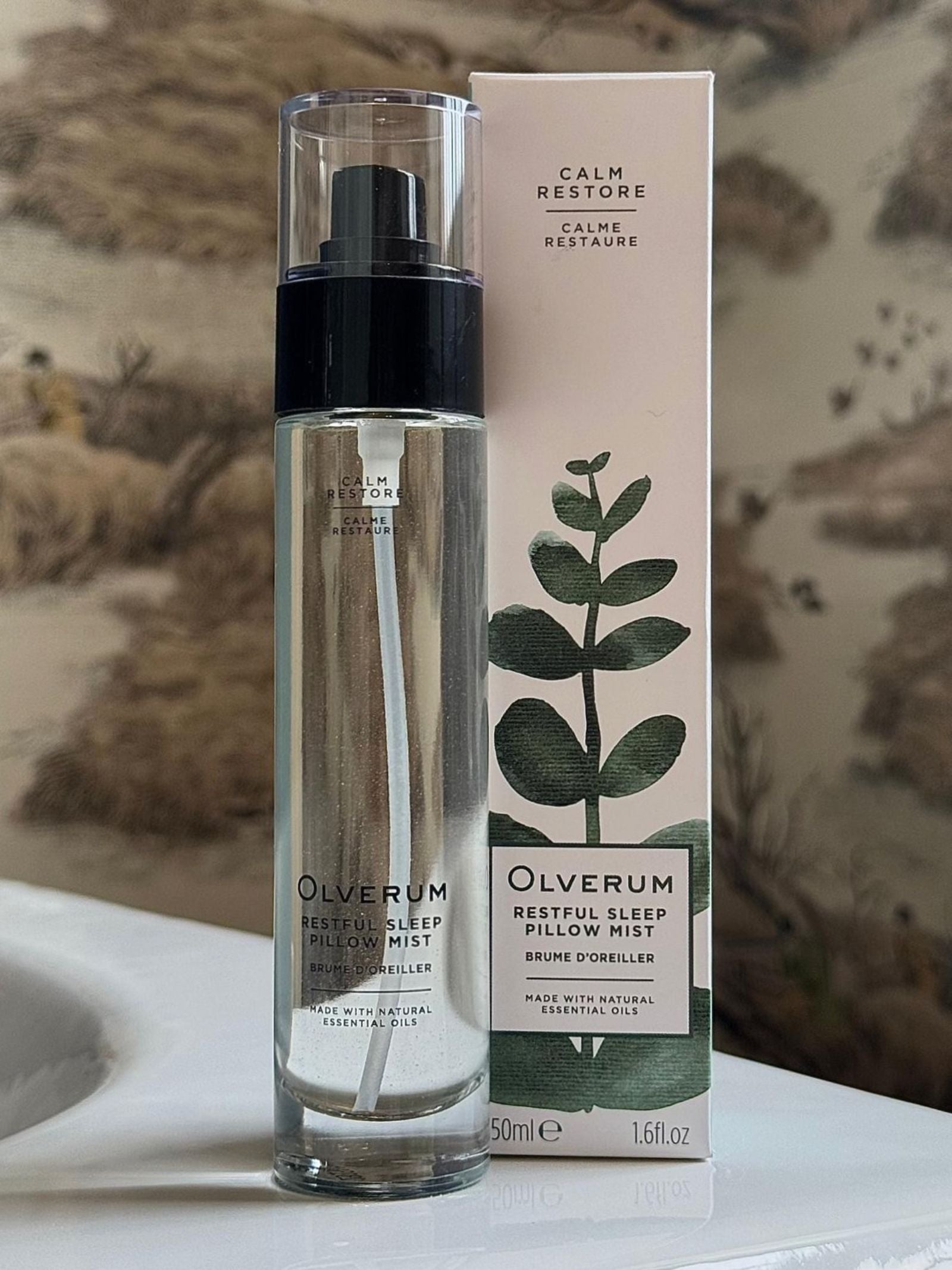 A bottle and box of OLVERUM Restful Sleep Pillow Mist are displayed on a white surface; the box shows leafy artwork and text, set against a softly blurred, nature-inspired background.