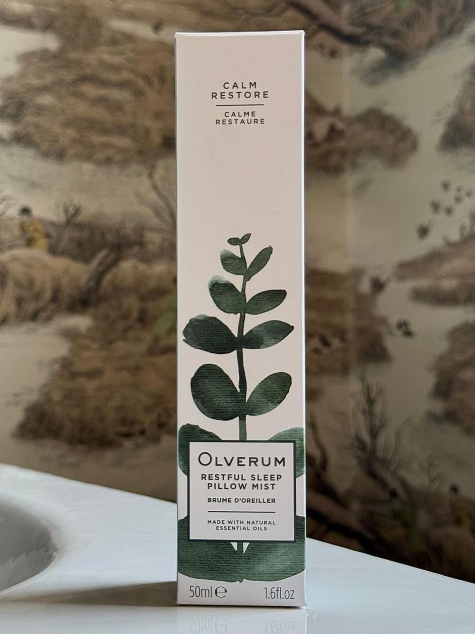 A box of OLVERUM Restful Sleep Pillow Mist stands upright on a white surface, featuring botanical packaging that highlights its calming aromatic benefits. Behind it, a wall mural with a nature theme completes the scene.