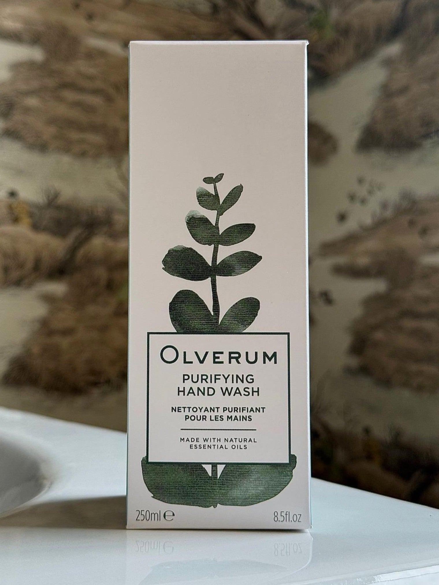 A box of OLVERUM Purifying Hand Wash, featuring a green botanical design on the front, stands on a white surface with an earthy-toned background. The packaging emphasizes its vegan-friendly formula and natural origin ingredients.