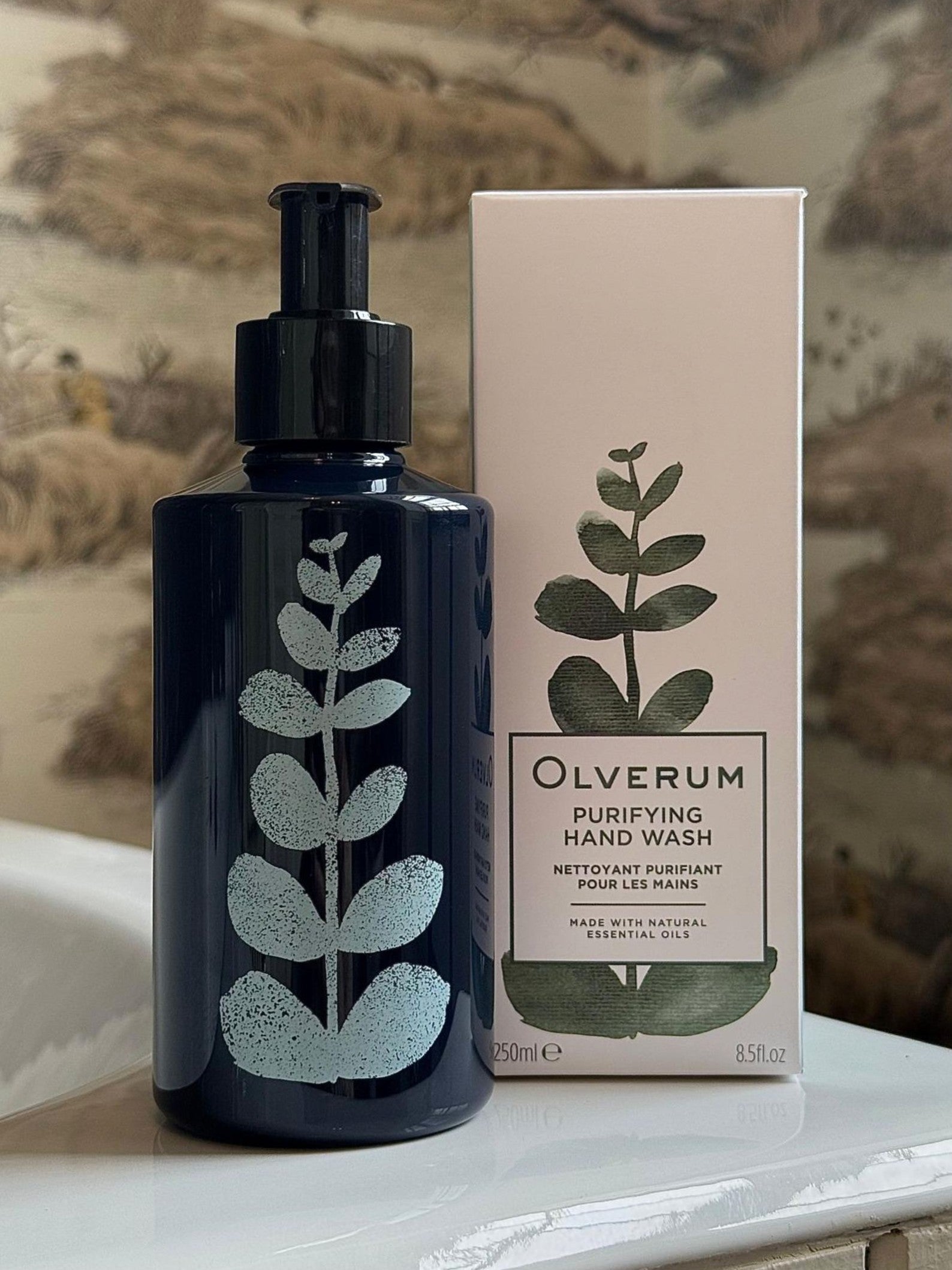 A dark bottle and white box of OLVERUM Purifying Hand Wash, featuring natural origin ingredients and a green leaf design, are shown on a bathroom counter in front of patterned wallpaper.