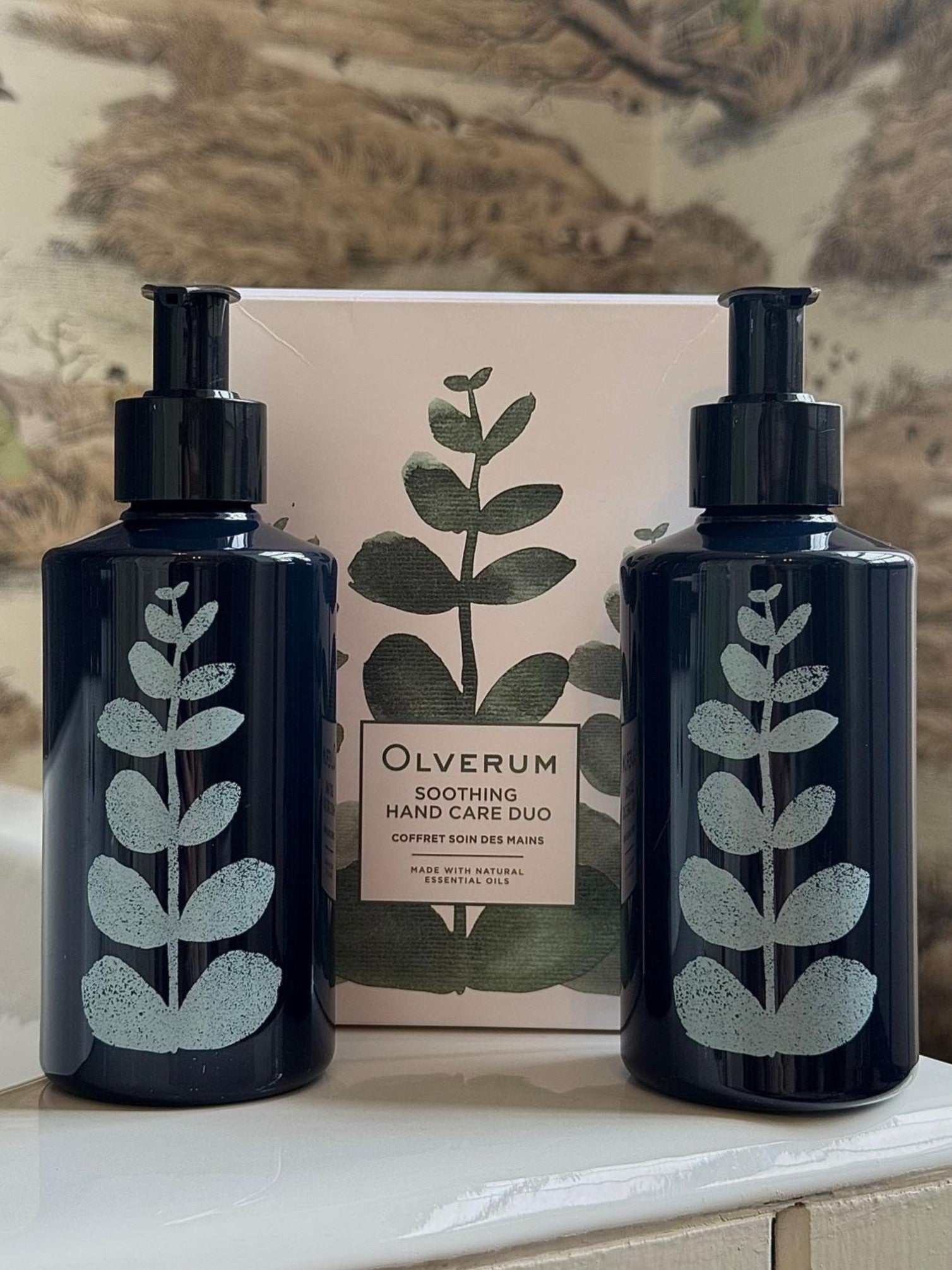 Two dark blue OLVERUM Hand Care Duo pump bottles with leaf motifs stand before a matching box against a neutral, botanical-patterned background.
