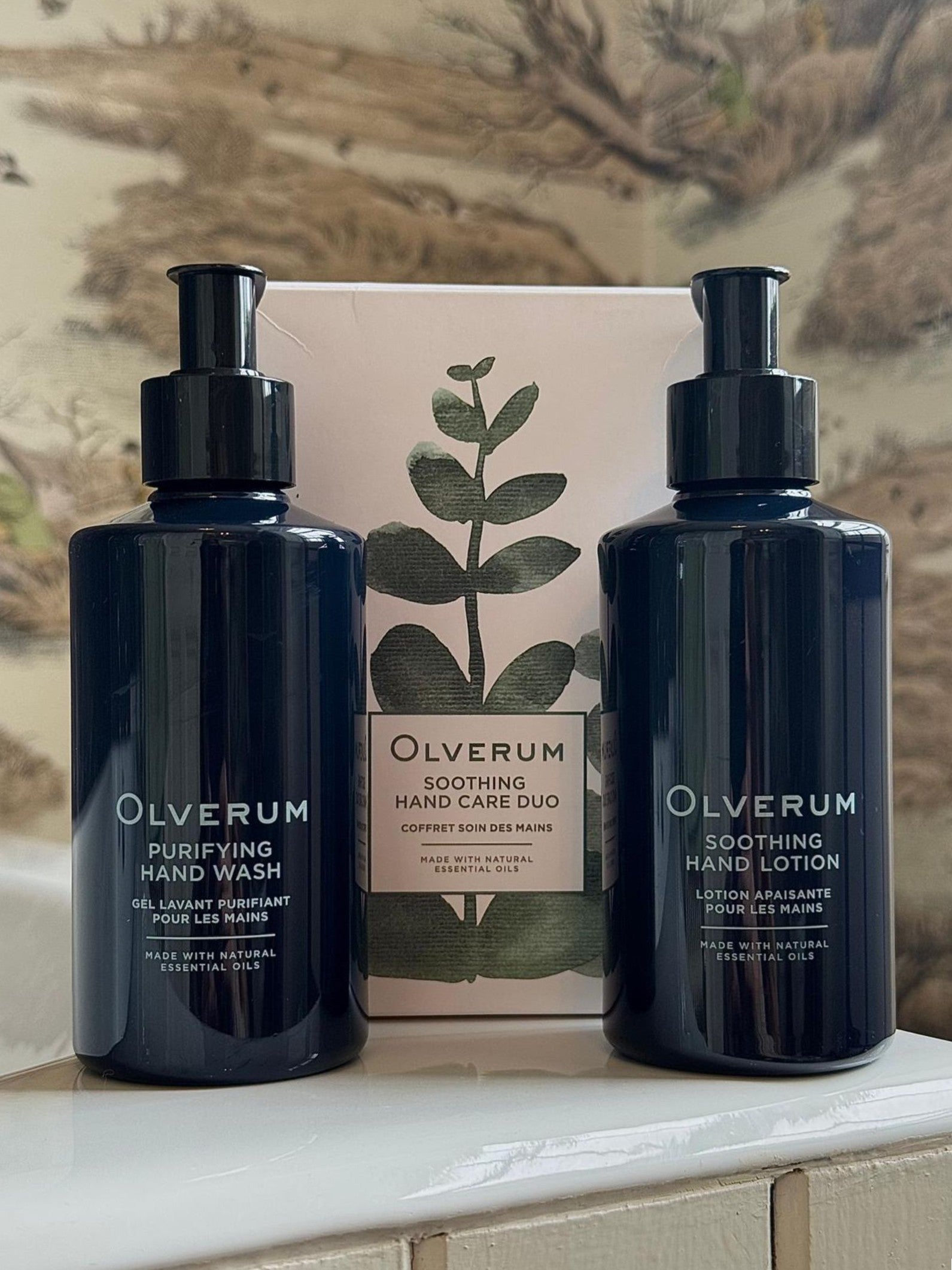 Two dark blue pump bottles labeled OLVERUM Purifying Hand Wash and Soothing Hand Lotion stand before a white box featuring a green plant illustration and the text "Olverum Hand Care Duo" from OLVERUM.
