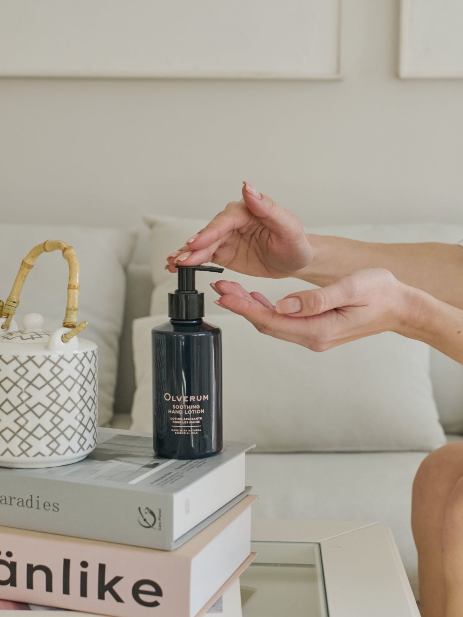 A person dispenses OLVERUM Soothing Hand Lotion onto their palm, with the bottle set on stacked books beside a bamboo-handled container on a coffee table. A neutral sofa appears in the background.