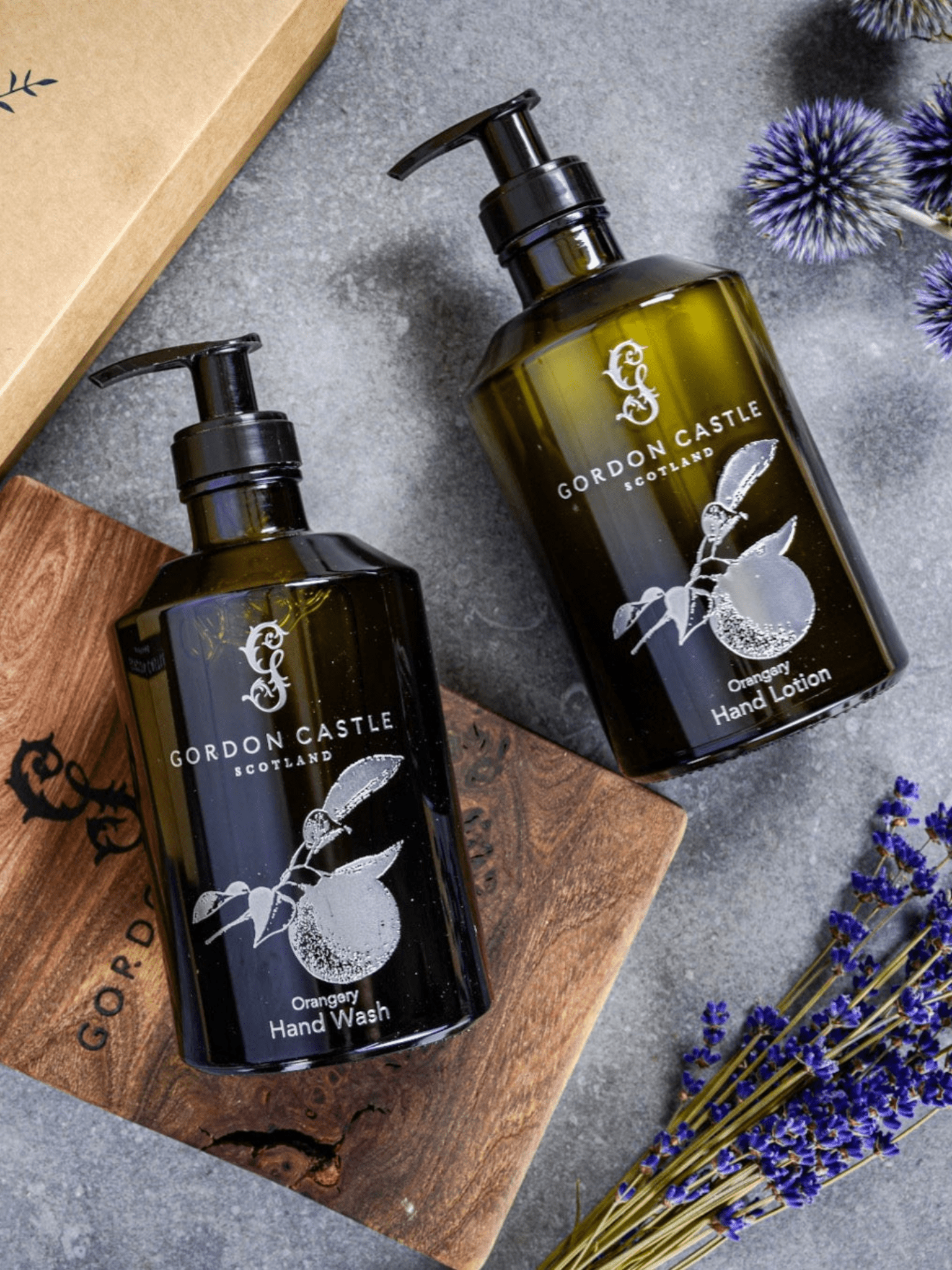 A Gordon Castle Orangery Hand Lotion in a refillable glass bottle is displayed on a wooden board with dried flowers and its box, creating an elegant scene. The lotion is infused with botanical extracts for a luxurious touch.