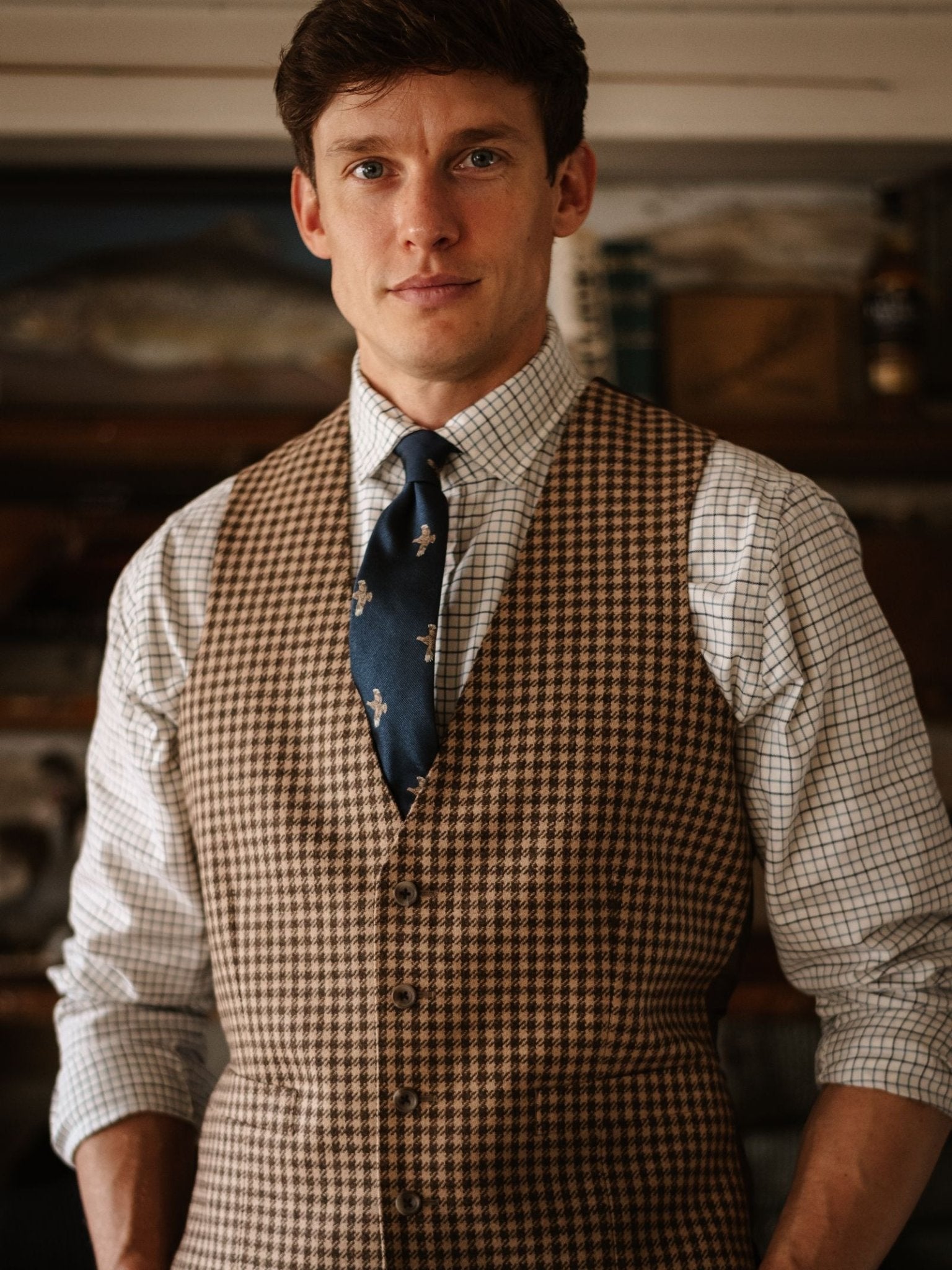 A man wears Campbell's of Beauly's Partridge Silk Tie, featuring a subtle partridge motif, paired with a brown checkered vest and plaid shirt, standing indoors with blurred shelves and books in the background for countryside charm.