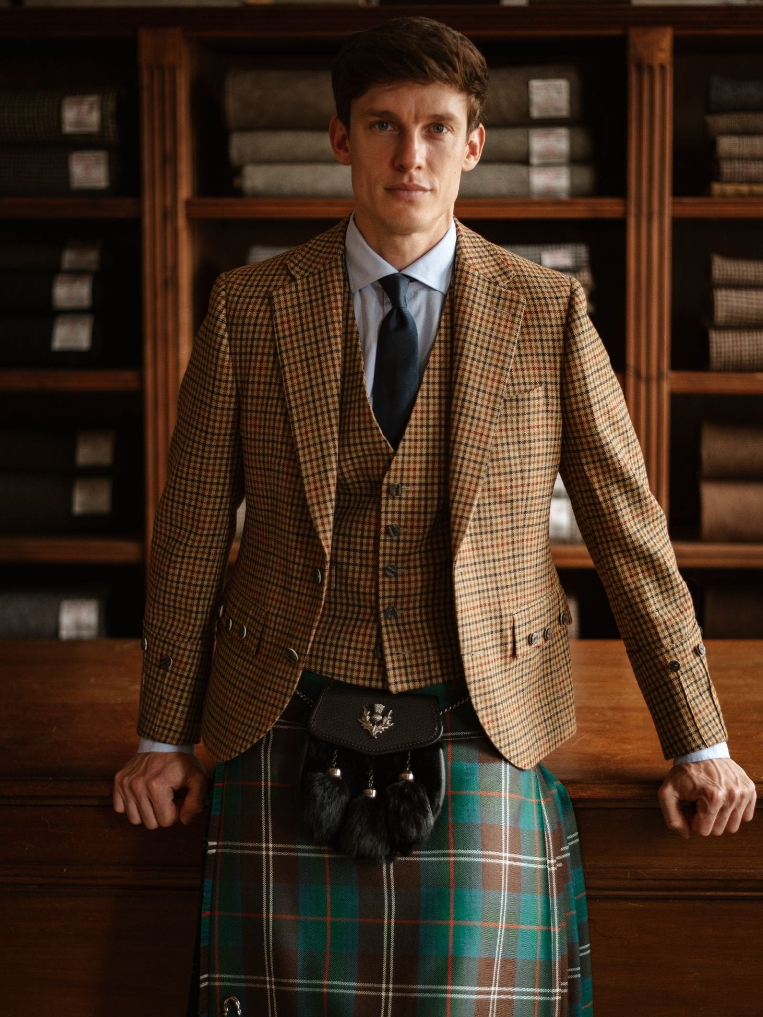 A man in a brown checked suit jacket and vest, light blue shirt, Plain Silk Tie by Campbell's of Beauly, and green tartan kilt with a black sporran stands indoors before wooden shelves stacked with folded fabric.
