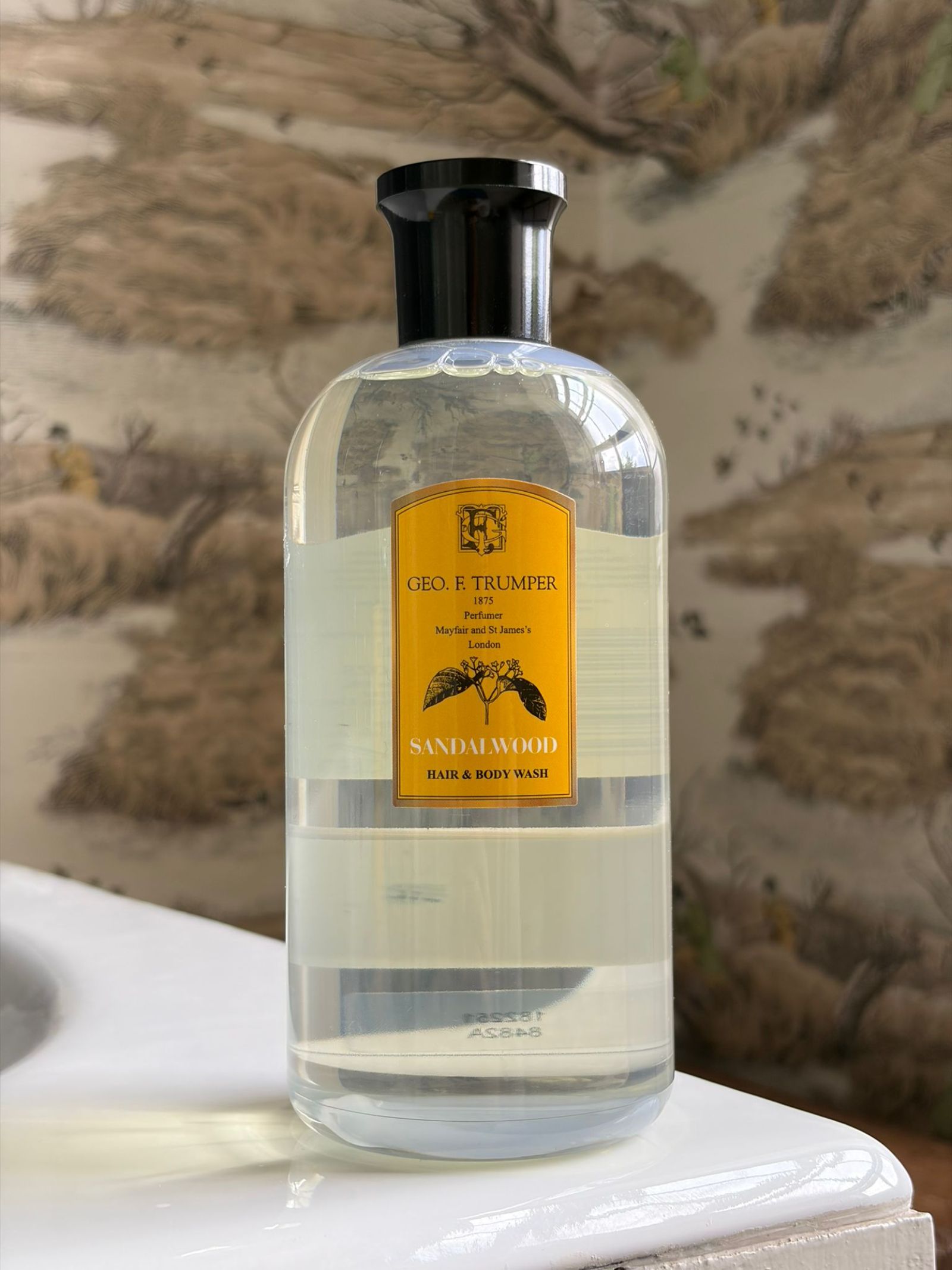 A clear bottle of Geo. F. Trumper Hair & Body Wash with a yellow label and black cap sits on a bathroom counter, its warm scent enhancing the natural-themed wallpaper in the background.