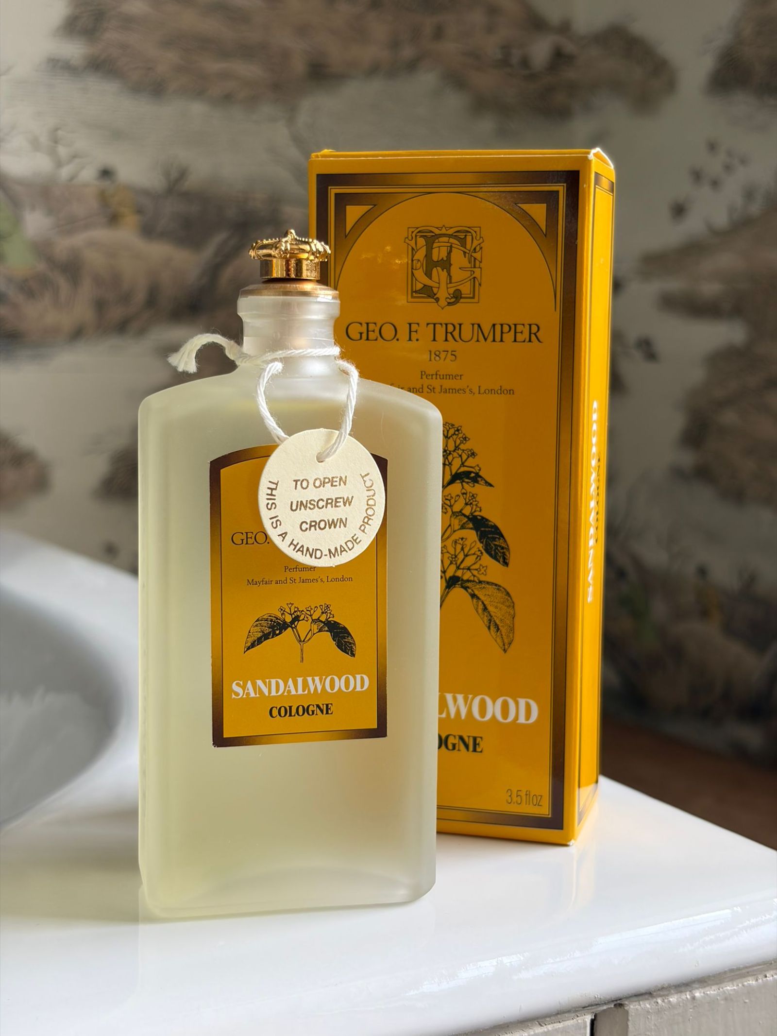 A frosted glass bottle of Geo. F. Trumper Sandalwood Cologne with a gold cap sits next to its yellow box. A round tag on the neck reads, "To open unscrew crown." Hand-made with Mysore sandalwood oil for a masculine scent.