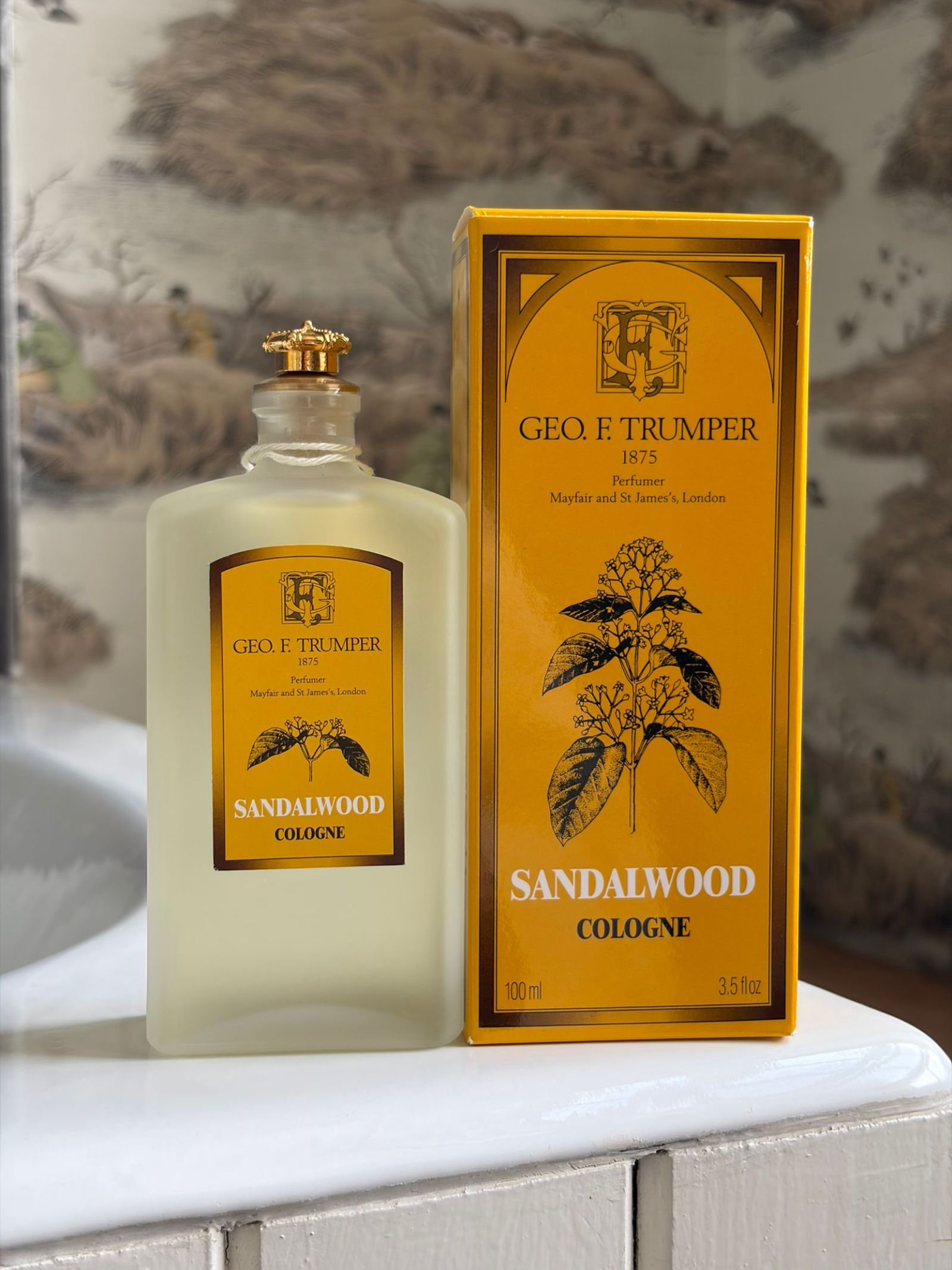 A bottle and box of Geo. F. Trumper Sandalwood Cologne rest on a white surface; the yellow box with black botanical art complements the frosted glass bottle’s gold cap and matching label.