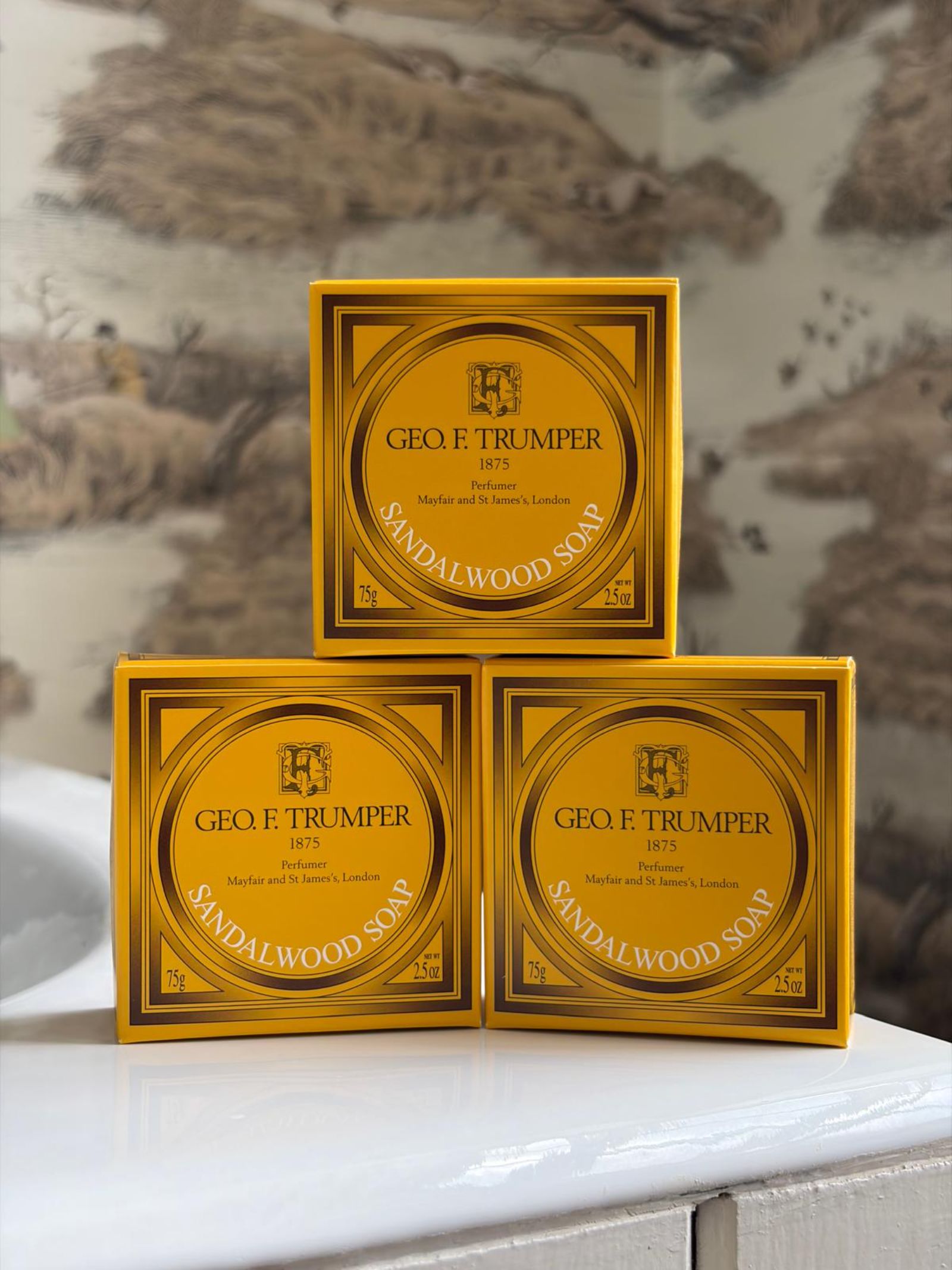 Three yellow boxes of Geo. F. Trumper Sandalwood Hand Soaps, Set of 3, are stacked in a pyramid on a white surface against blurred wallpaper—an elegant addition to any guest bathroom.