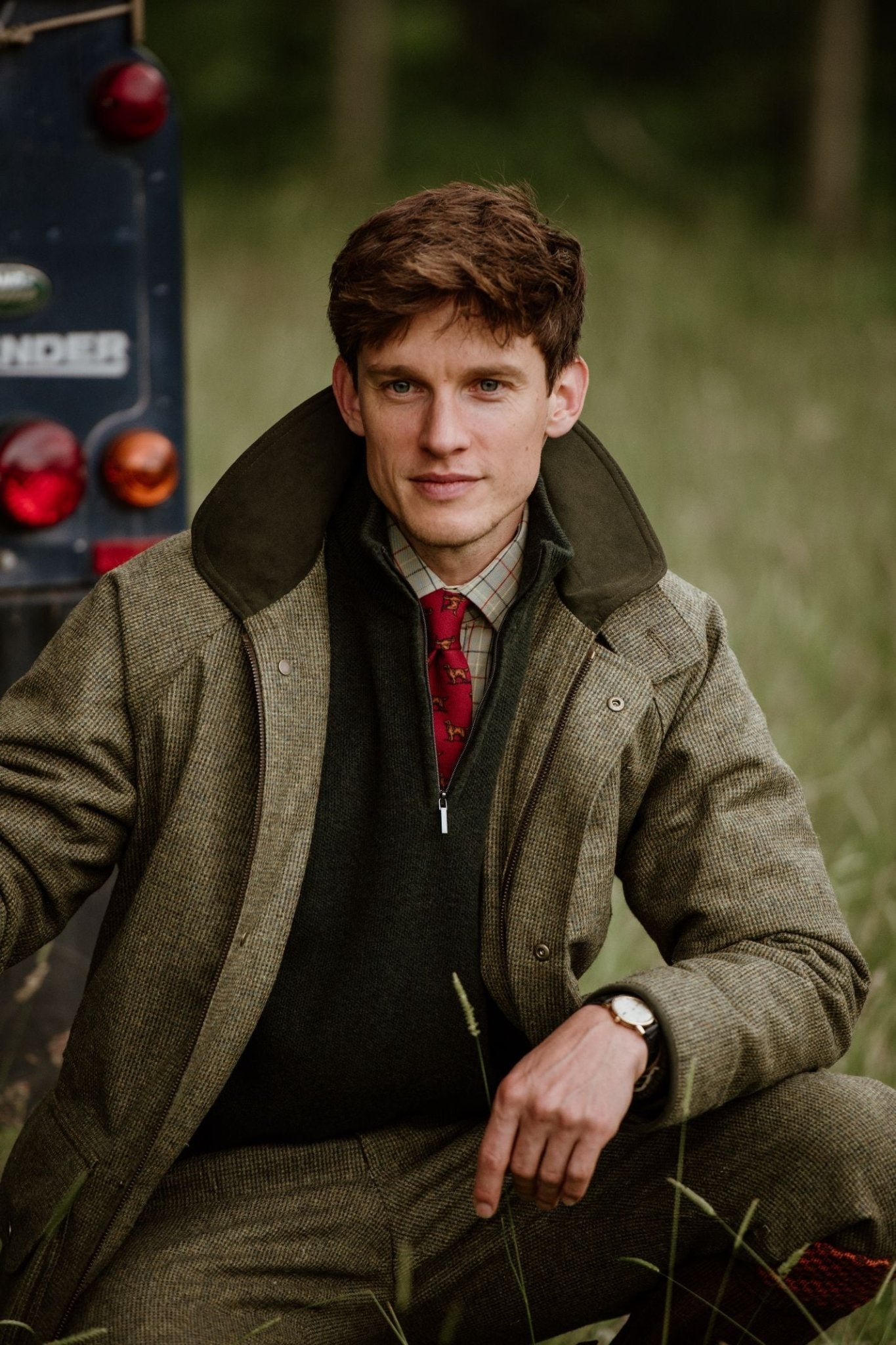 A man in tweed attire kneels next to a dark vehicle in a grassy field, wearing a tweed jacket, dark green jumper, collared shirt, and the Setter Dog Silk Tie by Campbell's of Beauly.