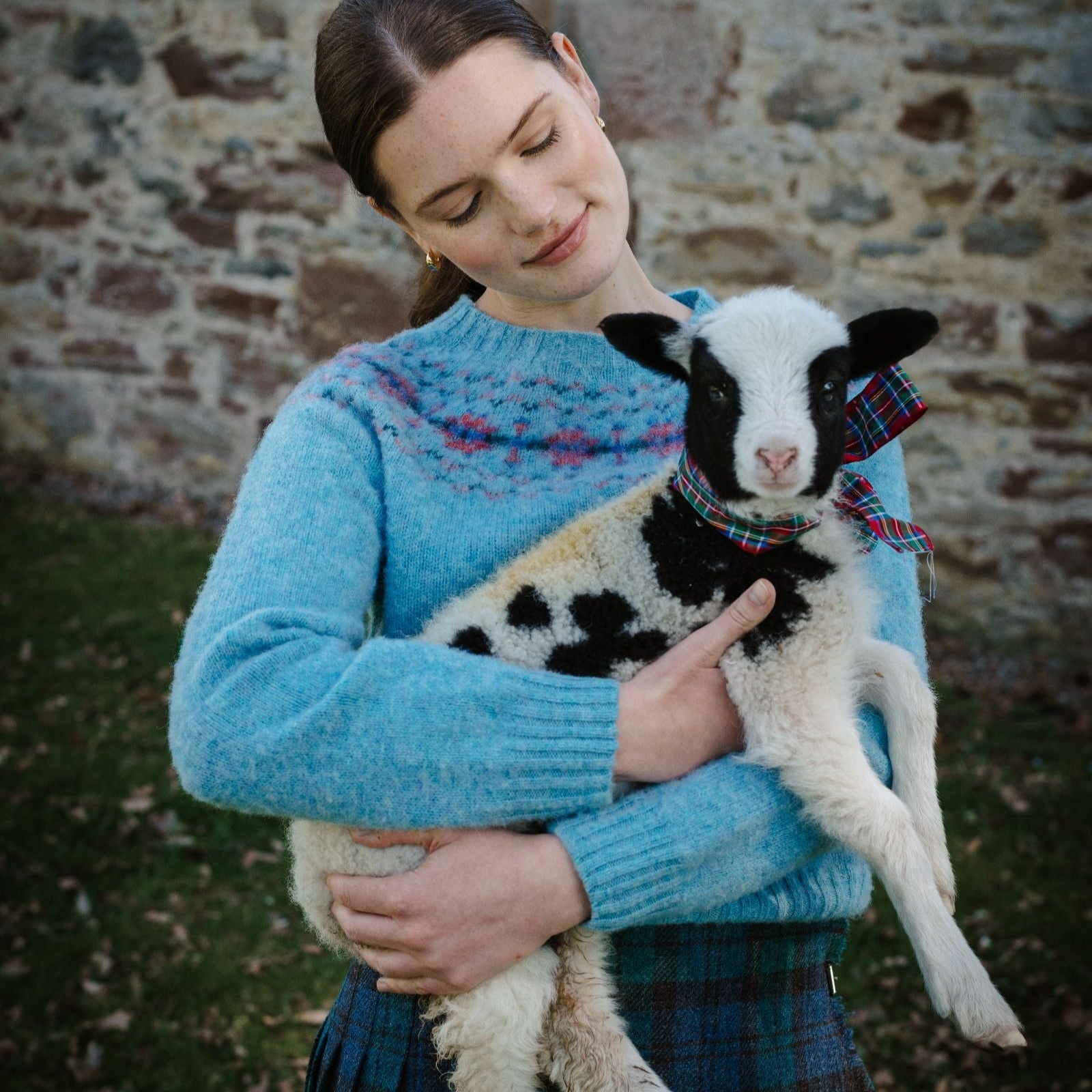 A woman in a blue sweater holds a black-and-white lamb wearing a tartan ribbon around its neck. They stand outside by a stone wall, and the woman looks affectionately at the lamb.