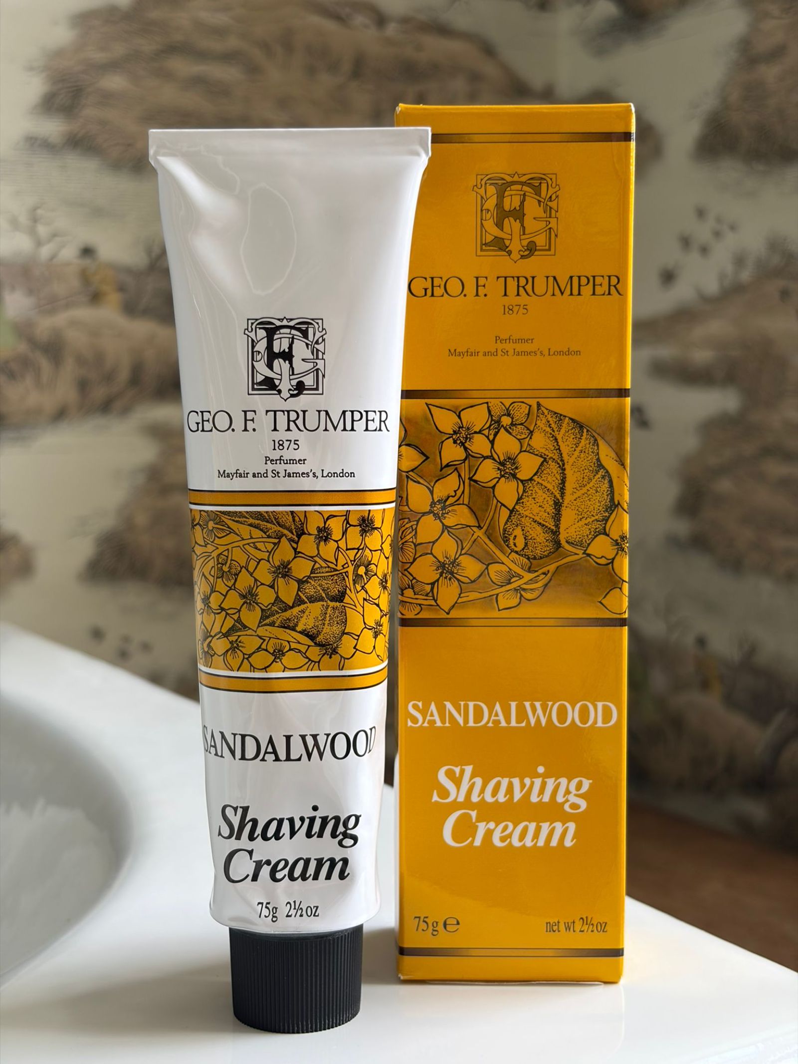 A tube and box of Geo. F. Trumper Sandalwood Soft Shaving Cream, ideal for sensitive skin, are on a bathroom counter. The white tube has black and gold accents; the yellow box features floral artwork.