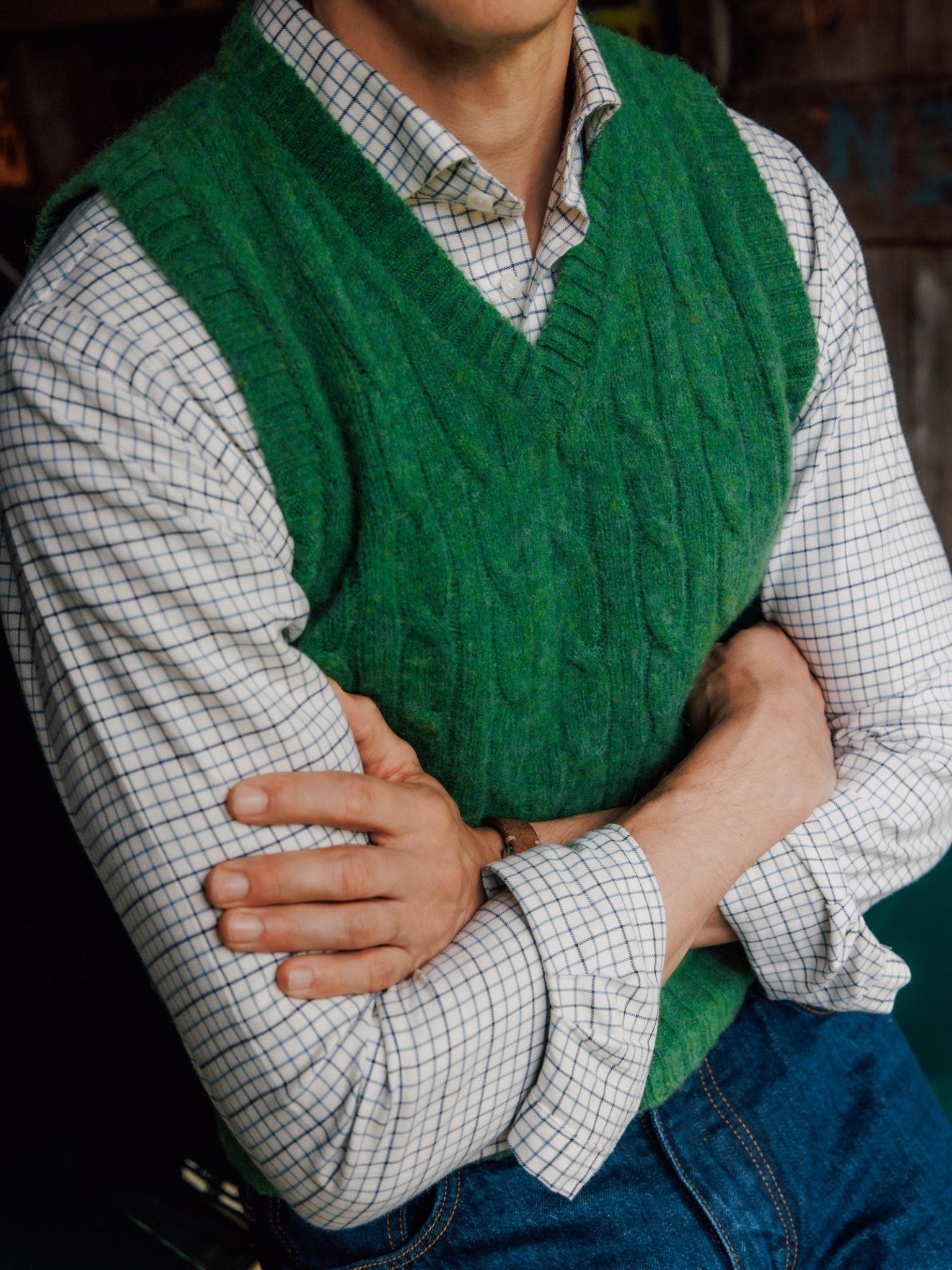 A person with arms crossed wears a green Campbell's of Beauly Shetland Cable Slipover over a white checkered shirt and blue jeans. The photo is cropped to focus on the torso and folded arms.