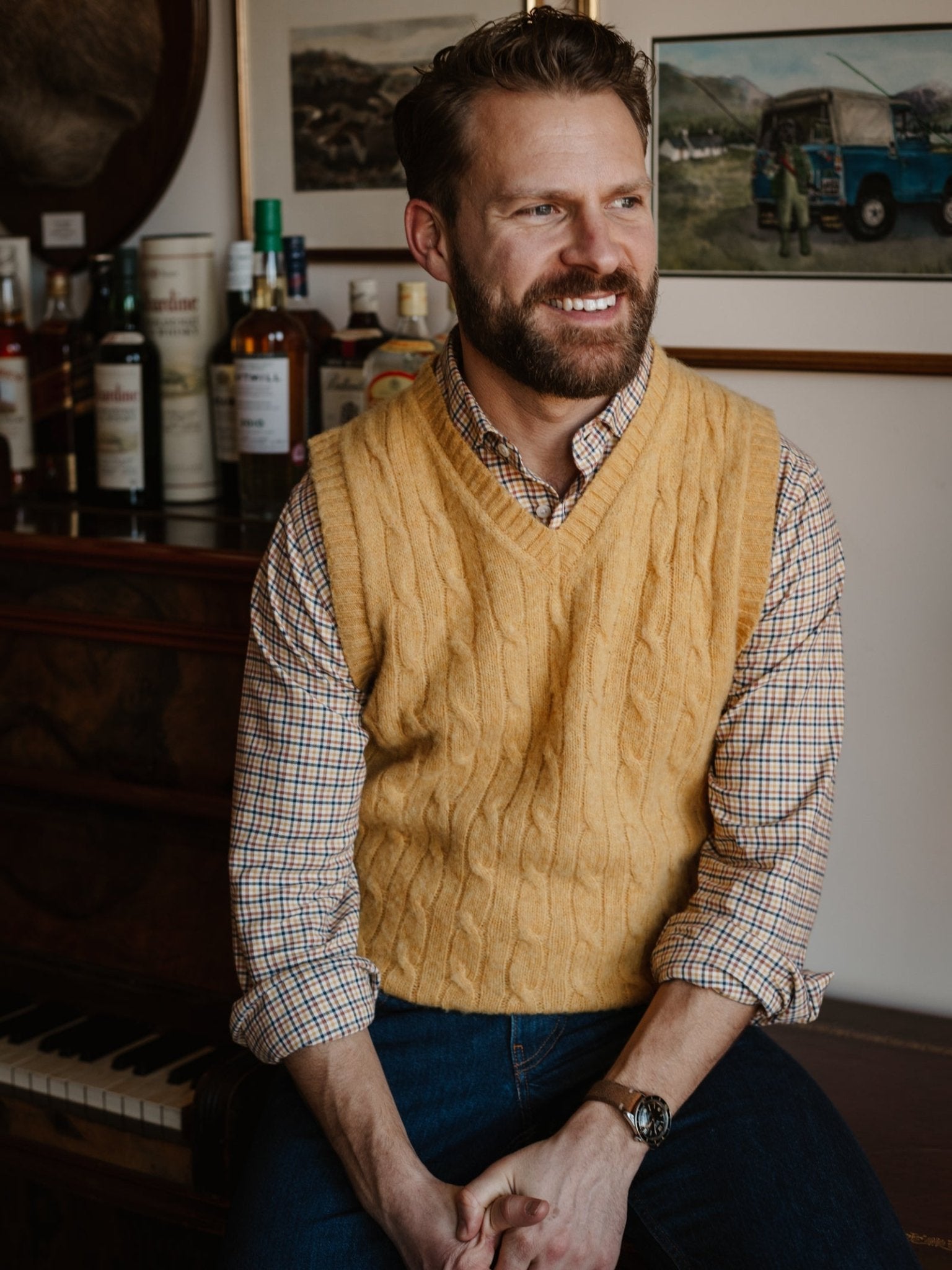 A smiling bearded man sits on a piano bench, wearing the Shetland Cable Slipover by Campbell's of Beauly over a plaid shirt and jeans. Bottles and framed pictures decorate the background.