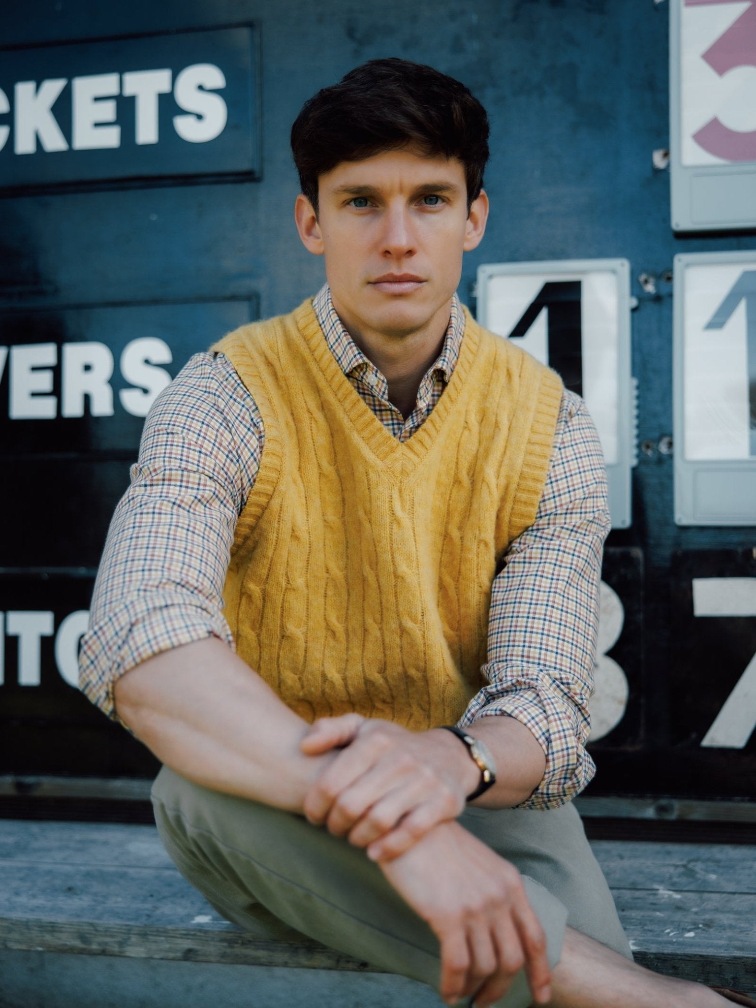 A man wearing the Campbell's of Beauly Shetland Cable Slipover over a plaid shirt sits on steps in front of a scoreboard, gazing at the camera with his arms on his knees.