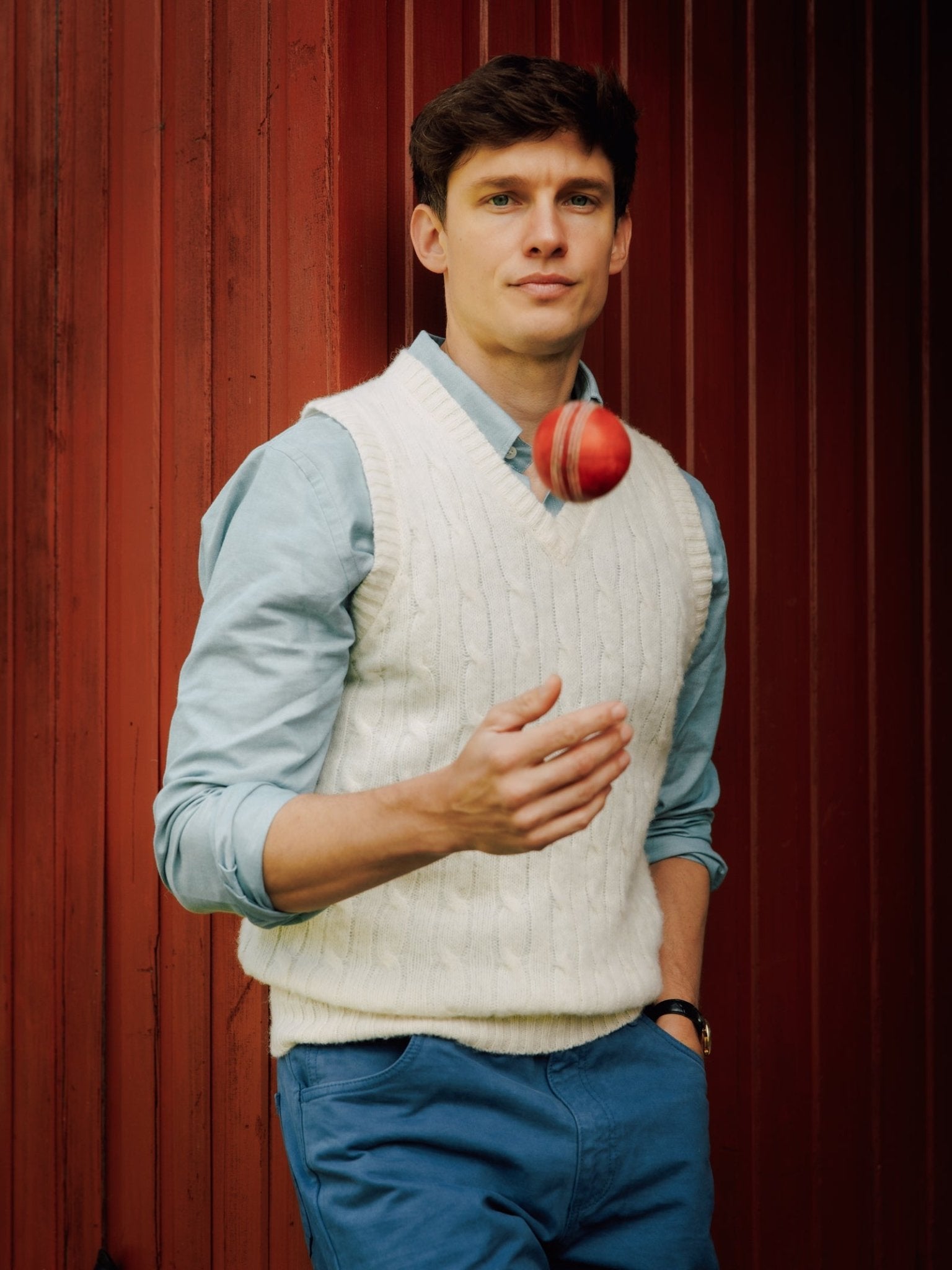 A young man in a Campbell's of Beauly Shetland Cable Slipover and blue shirt tosses a red cricket ball in the air, standing against a red wooden wall.