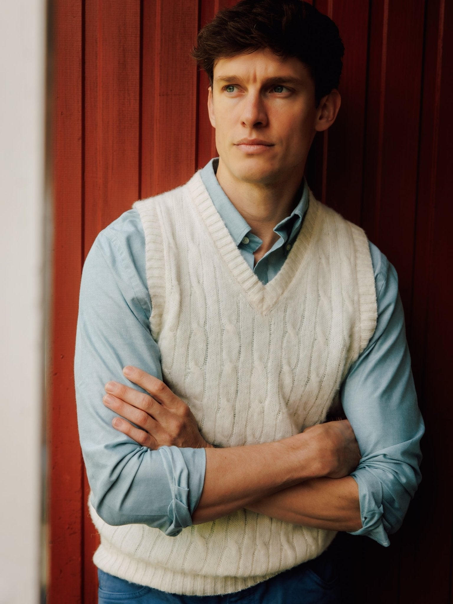 A young man with short brown hair stands in front of a red wooden wall, wearing a light blue shirt and the Shetland Cable Slipover by Campbell's of Beauly. With arms crossed, he looks thoughtfully into the distance.