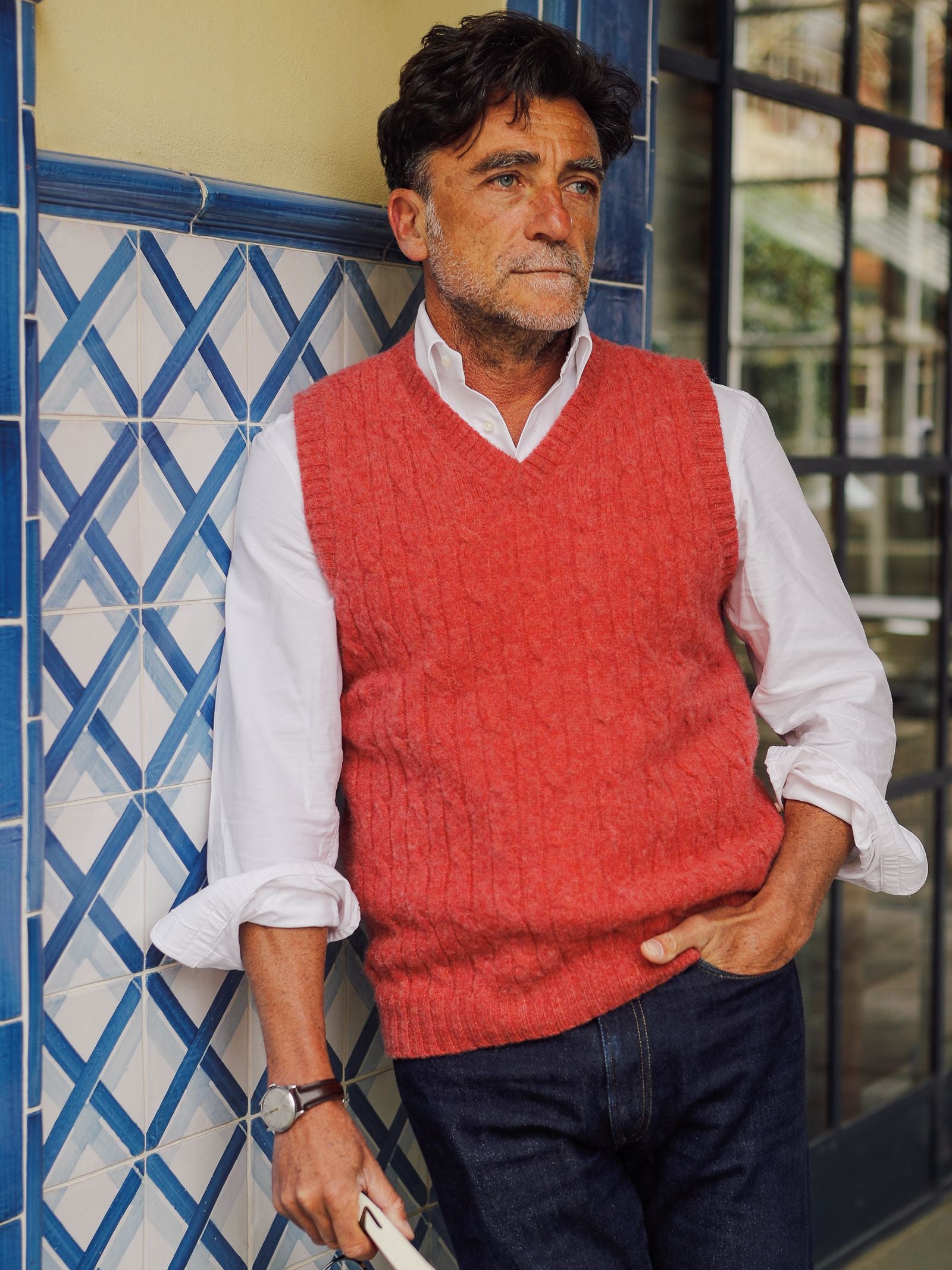 A man in a red Campbell’s of Beauly Shetland Cable Slipover layered over a white shirt and dark jeans stands beside a blue-tiled wall, holding a book with one hand in his pocket, gazing thoughtfully into the distance.