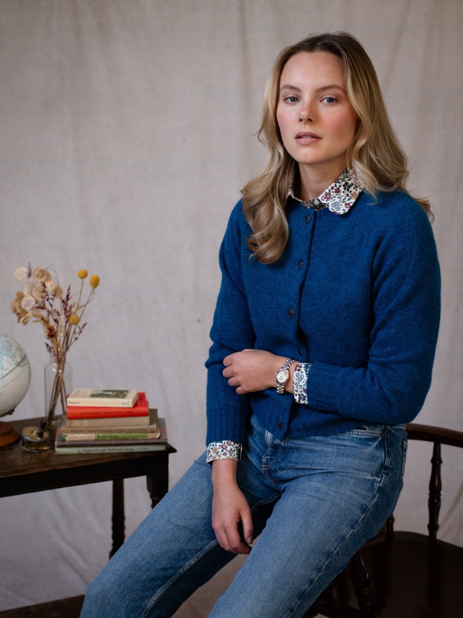 A woman with blonde hair sits on a chair, wearing Campbell's of Beauly Shetland Cropped Cardigan in blue over a floral-collared shirt and jeans. Books, a globe, and dried flowers rest on a nearby table against a plain neutral background.