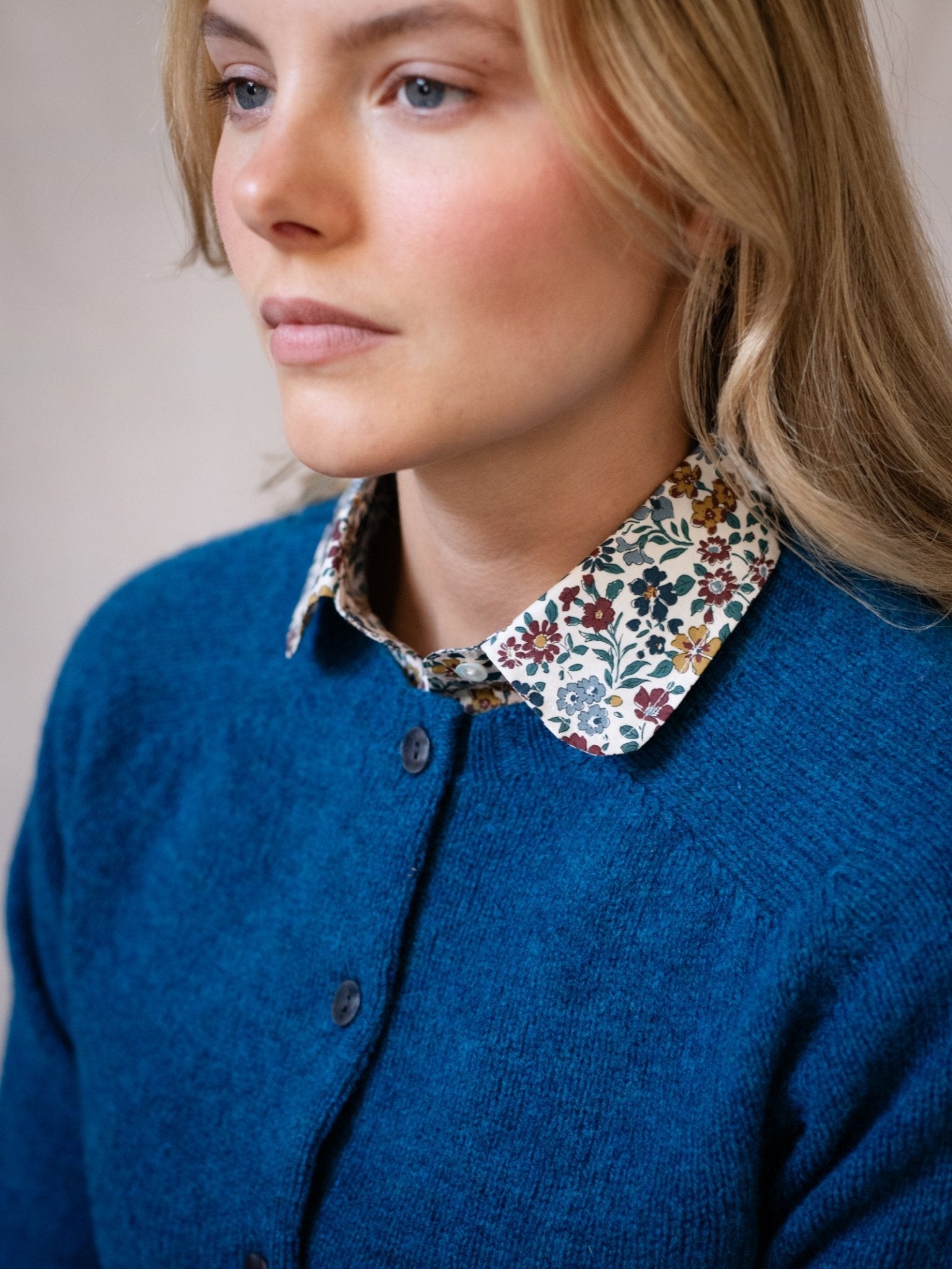 A woman with blonde hair wears a blue Shetland Cropped Cardigan by Campbell's of Beauly over a floral collared shirt, looking slightly to the side with a neutral expression.