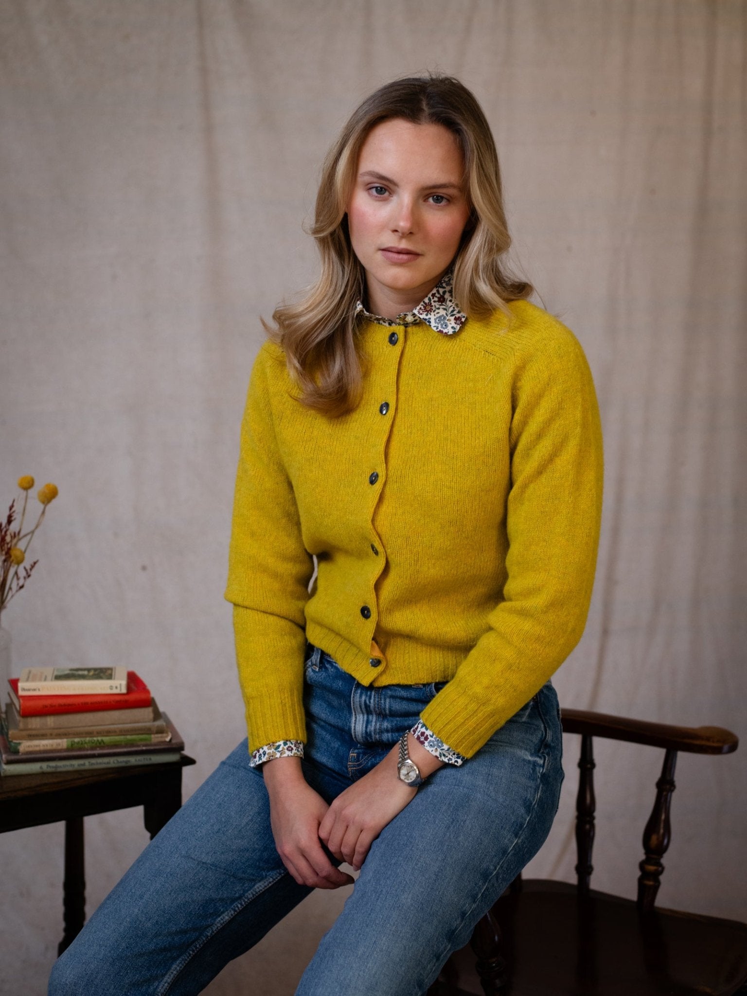 A blonde woman in a yellow Shetland Cropped Cardigan by Campbell's of Beauly and jeans sits on a wooden chair beside a small table with books and flowers, against a plain, light-colored fabric backdrop.