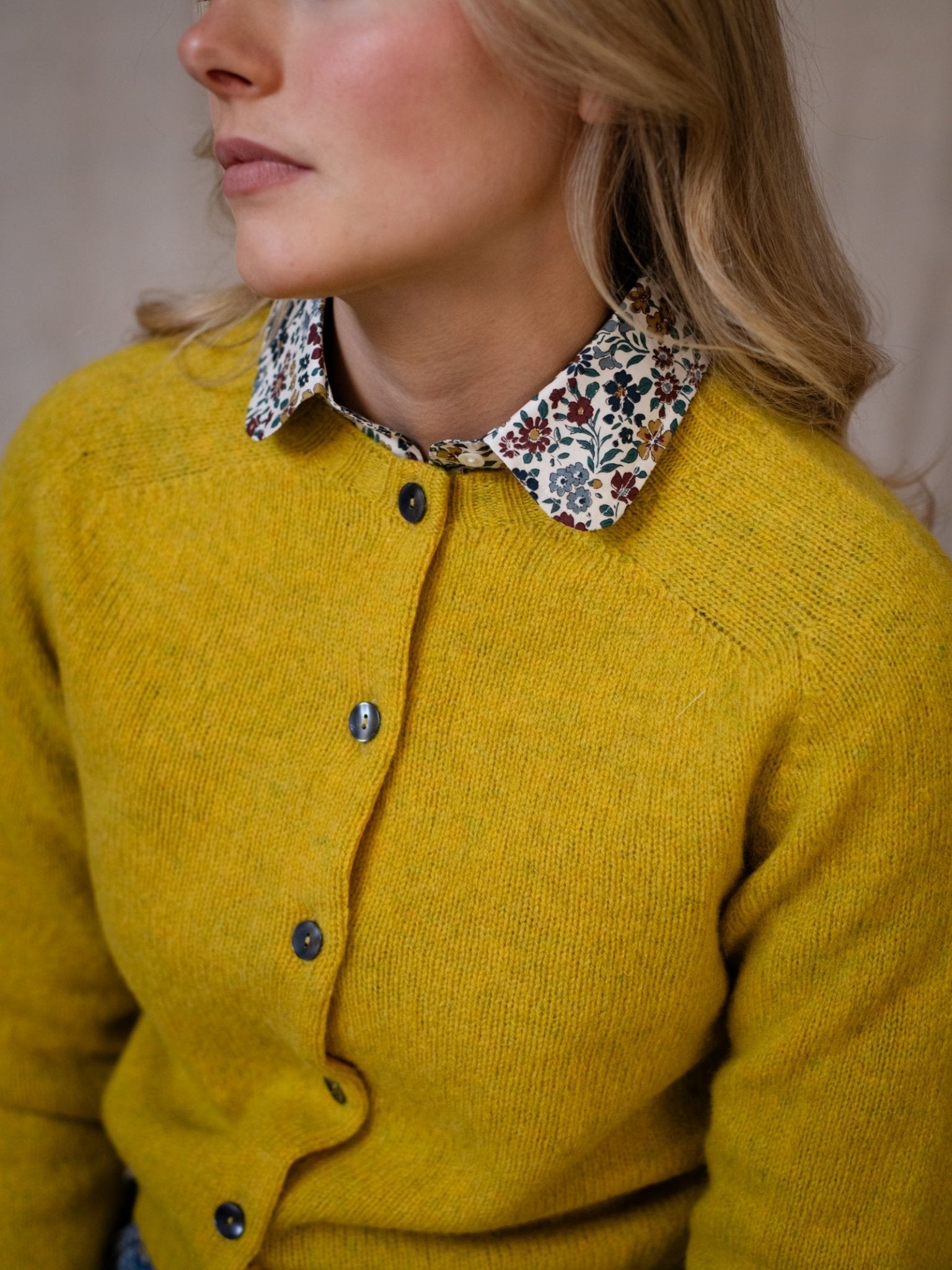A woman with long blonde hair wears a mustard yellow Shetland Cropped Cardigan from Campbell's of Beauly over a white shirt with a colorful floral collar. Facing slightly left, only her upper body and lower face are visible.