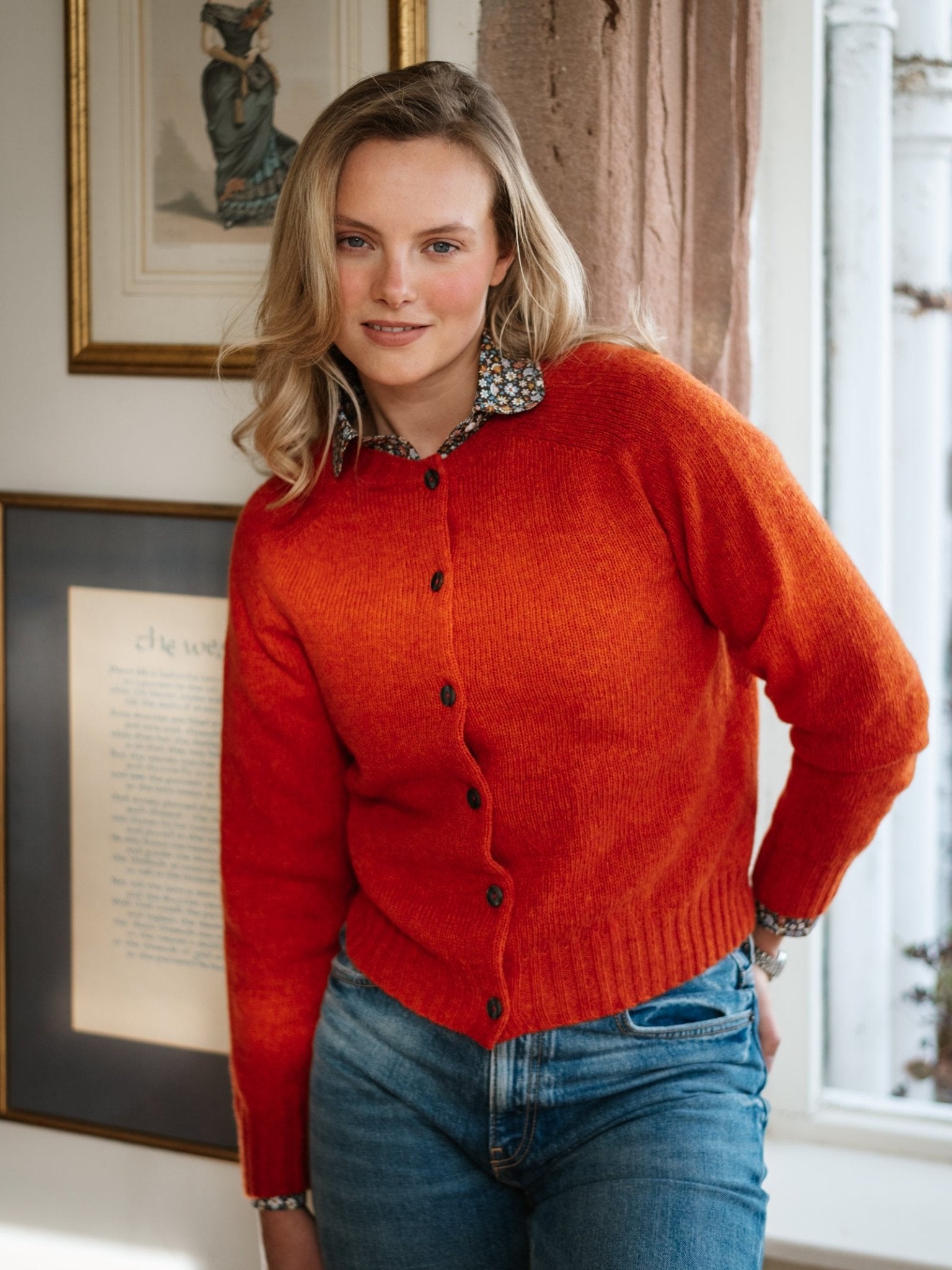 A woman with blonde hair wears a bright orange Shetland Cropped Cardigan by Campbell's of Beauly, made from Pure New Wool, paired with blue jeans. Indoors, she smiles softly; framed art and a window accent the quality Scottish knitwear.