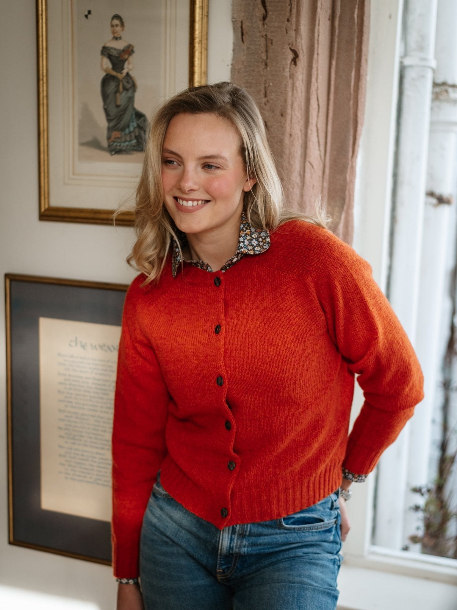 A woman with blonde hair smiles indoors in a bright orange Shetland Cropped Cardigan by Campbell's of Beauly and blue jeans. Framed art and sunlight from a window highlight the classic Scottish knitwear style.