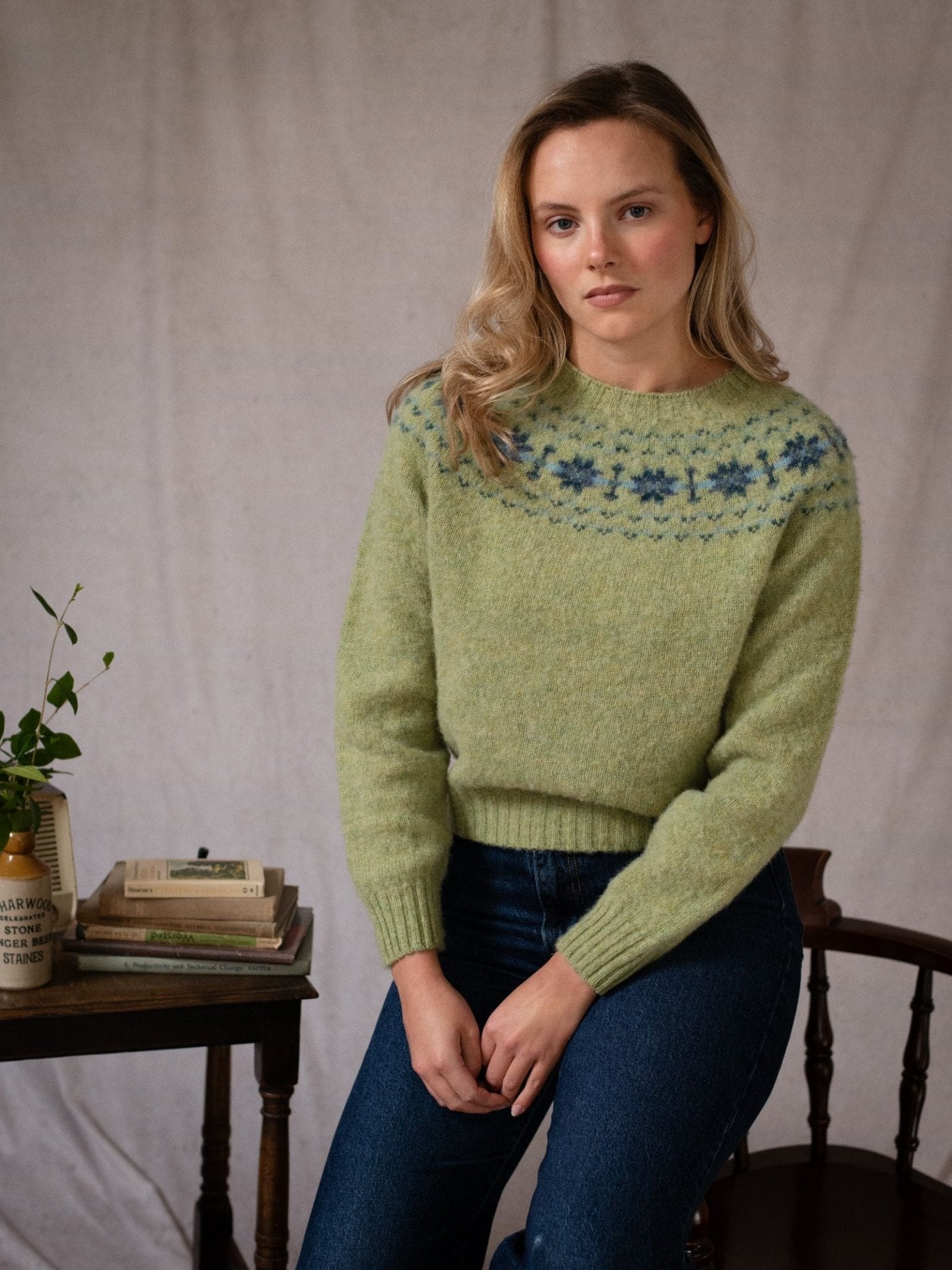 A person with long blonde hair wears the Shaggy Cropped Fairisle Crew Jumper by Campbell's of Beauly and blue jeans, sitting on a wooden chair next to a small table with books, a vase, and a plant against a neutral backdrop.