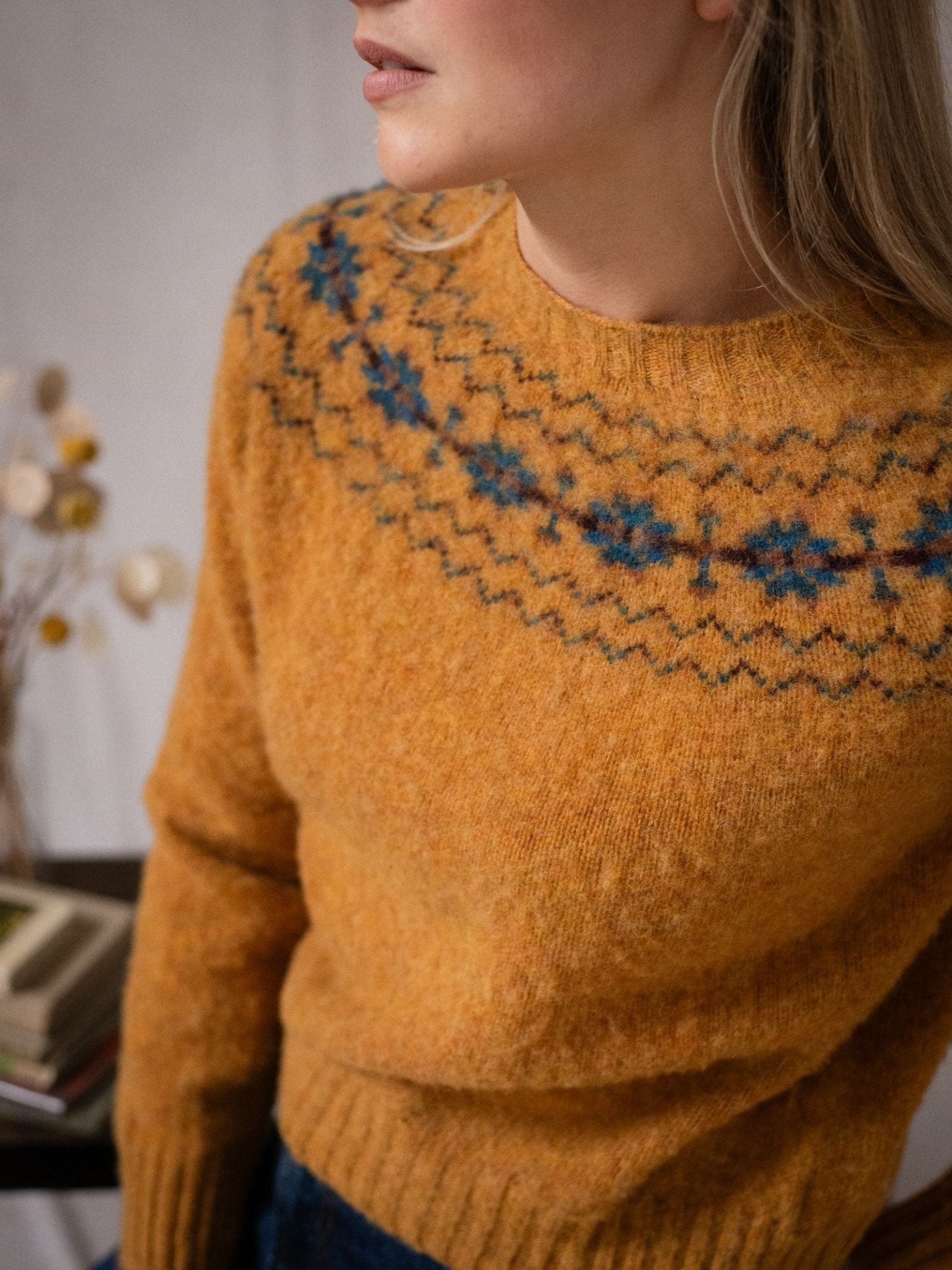 A woman with long blonde hair wears the Campbell's of Beauly Shaggy Cropped Fairisle Crew Jumper in orange Pure New Wool, featuring a blue yoke pattern. Books and dried flowers are seen in the background.