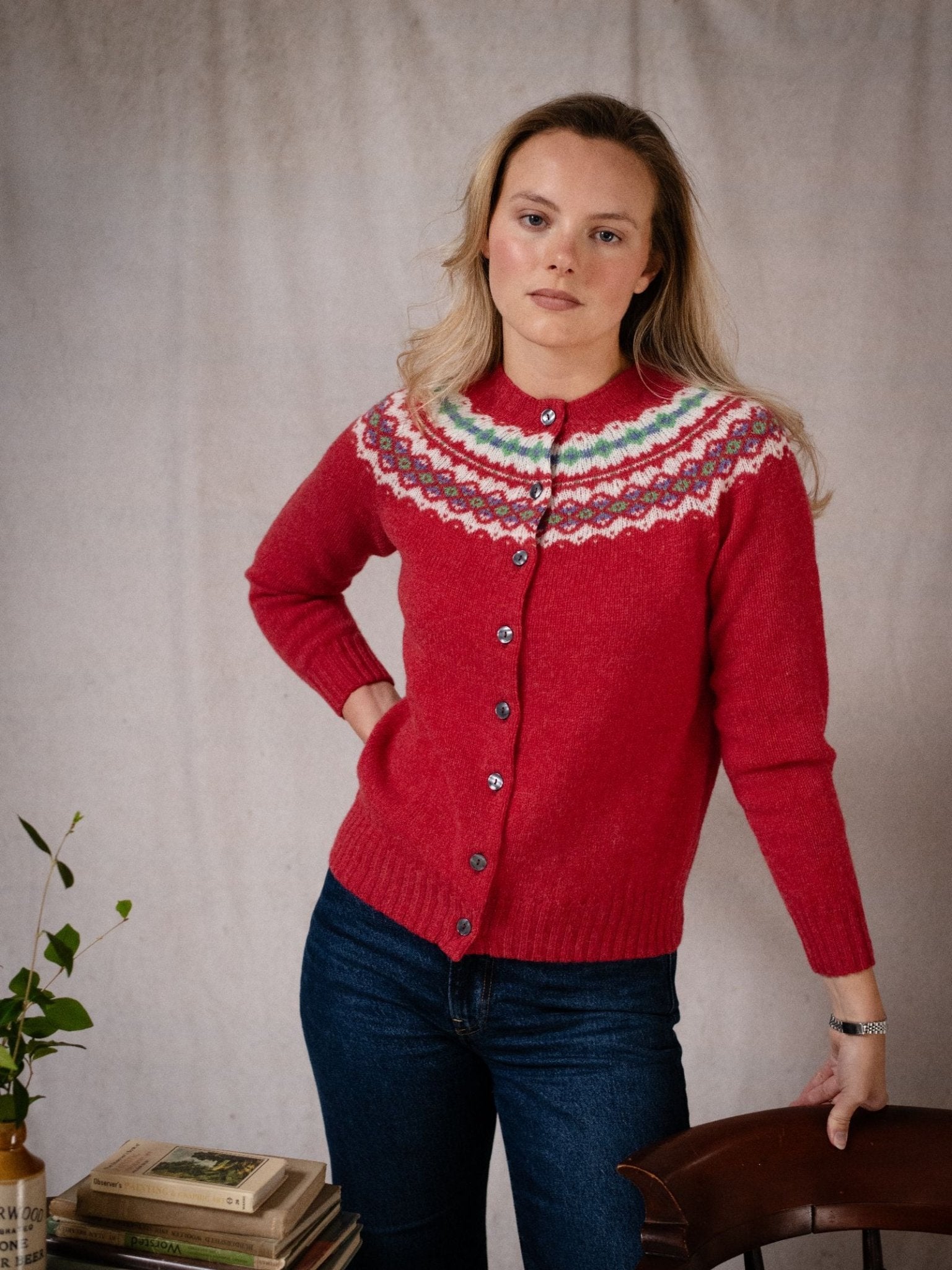 A blonde woman wears the Shetland Fairisle Cardigan from Campbell's of Beauly and blue jeans, standing indoors beside a wooden chair and a small table with stacked books and a vase of green leaves. She looks at the camera with a neutral expression.