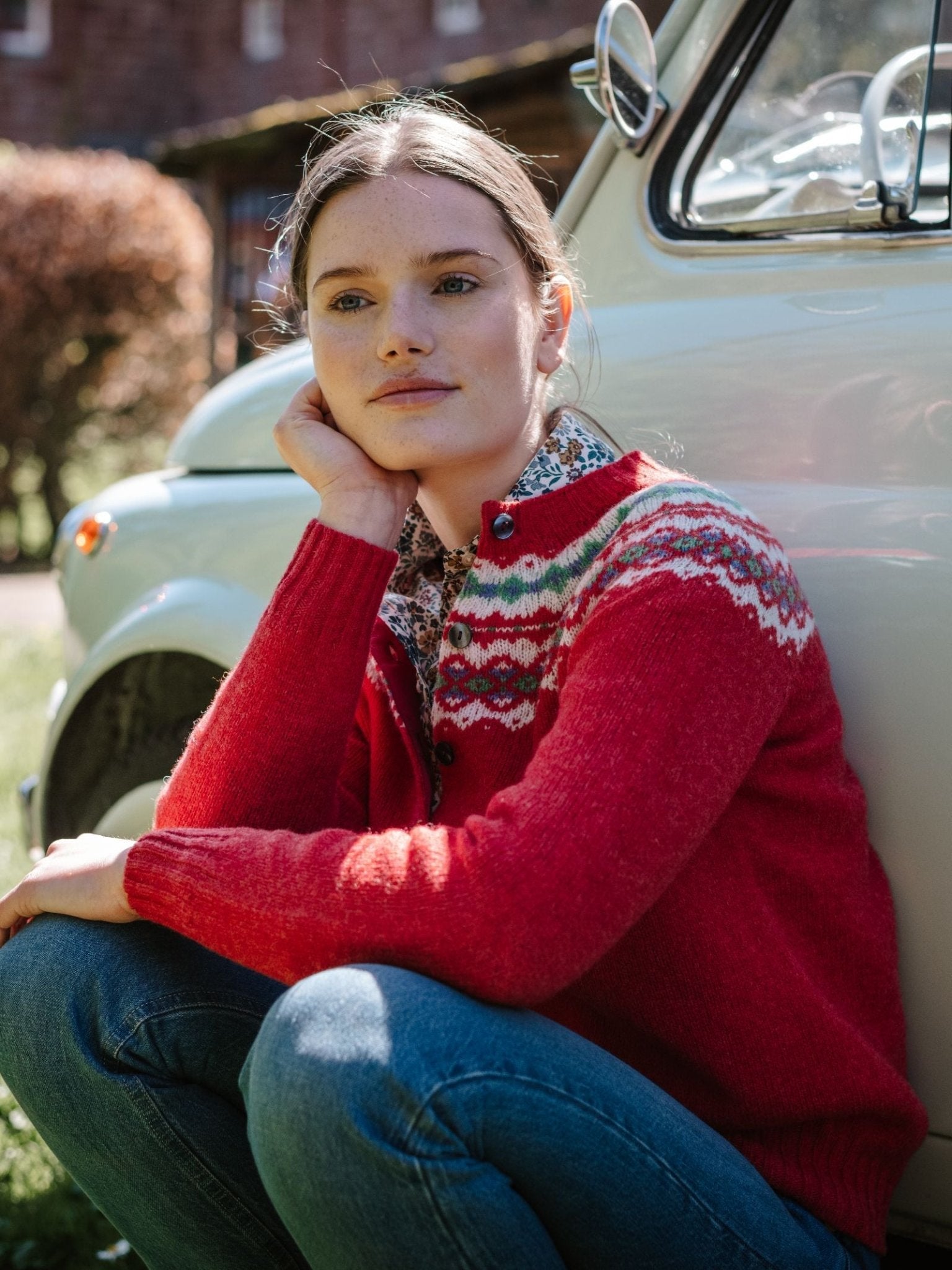 A woman in a Campbell's of Beauly Shetland Fairisle Cardigan and jeans sits outdoors by a vintage white car, resting her chin on her hand and gazing thoughtfully into the distance.