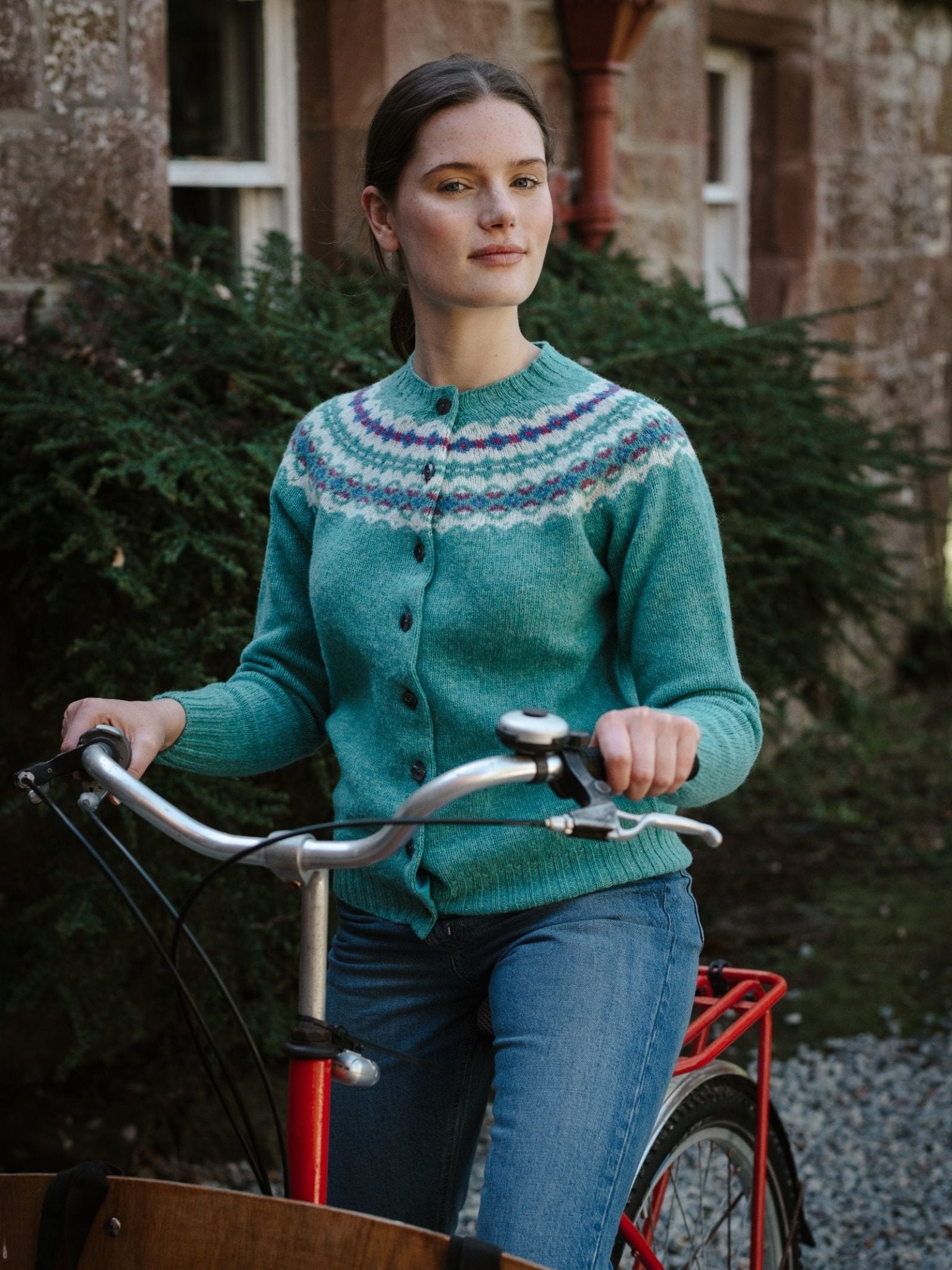A woman in a turquoise patterned Shetland Fairisle Cardigan by Campbell's of Beauly and blue jeans stands outside a brick building with greenery, holding the handlebars of a red bicycle.
